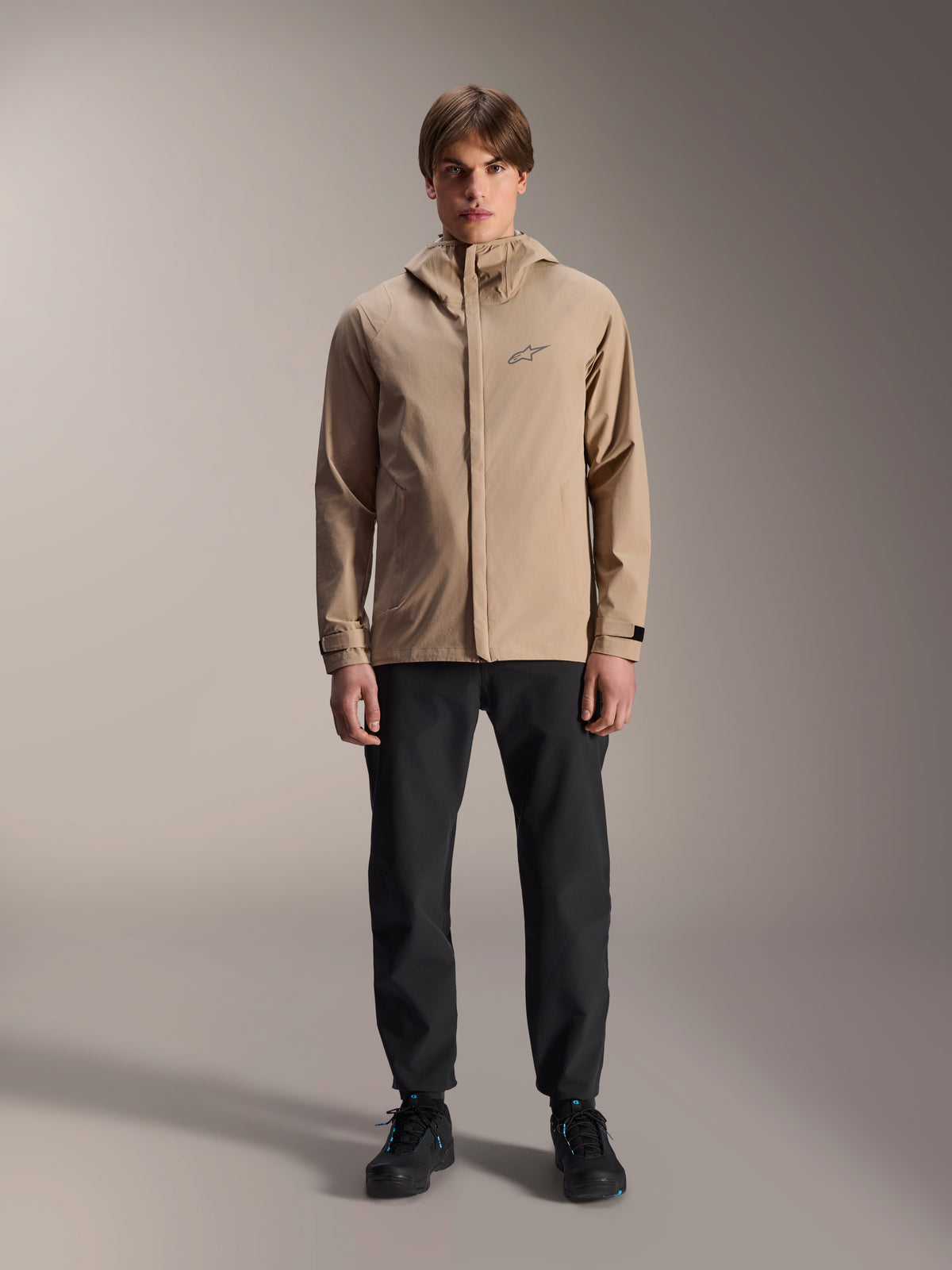 Alpinestars A-Dura Regenjacke, hellbraune Fahrradjacke, kombiniert mit schwarzer Hose und schwarzen Schuhen, mit kleinem Logo auf der Brust und Kapuze, professionelle Mountainbike-Bekleidung für Fahrten bei jedem Wetter.