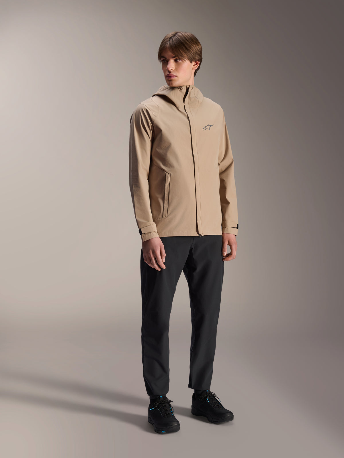 Männliches Model in der Alpinestars A-Dura Regenjacke in Hellbraun, kombiniert mit schwarzer Hose und Schuhen, minimalistische Outdoor-Mountainbike-Regenjacke mit Kapuzendesign und kleinem Logo auf der Brust.