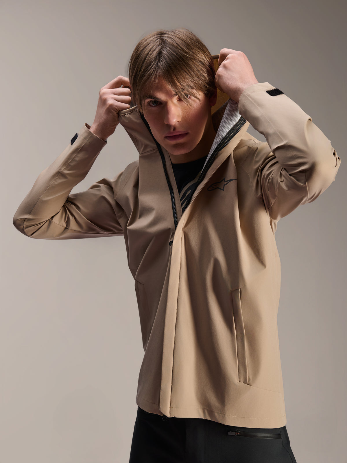Alpinestars A-Dura Regenjacke, MTB-Jacke, Hellbraun, Modell trägt die Jacke und setzt die elastische Kapuze auf, mit schwarzem Brustlogo, Reißverschlusstaschen und technischem, wasserdichtem 2,5-Lagen-Gewebe