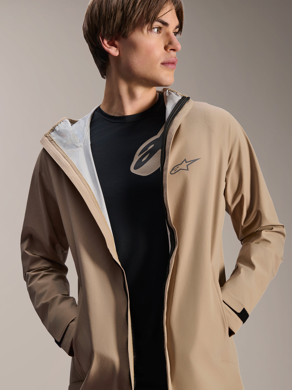 Männliches Model trägt die Alpinestars A-Dura Regenjacke, Hellbraun, geöffnet über einem schwarzen T-Shirt, weißes Mesh-Futter sichtbar, vielseitige wetterfeste Radjacke