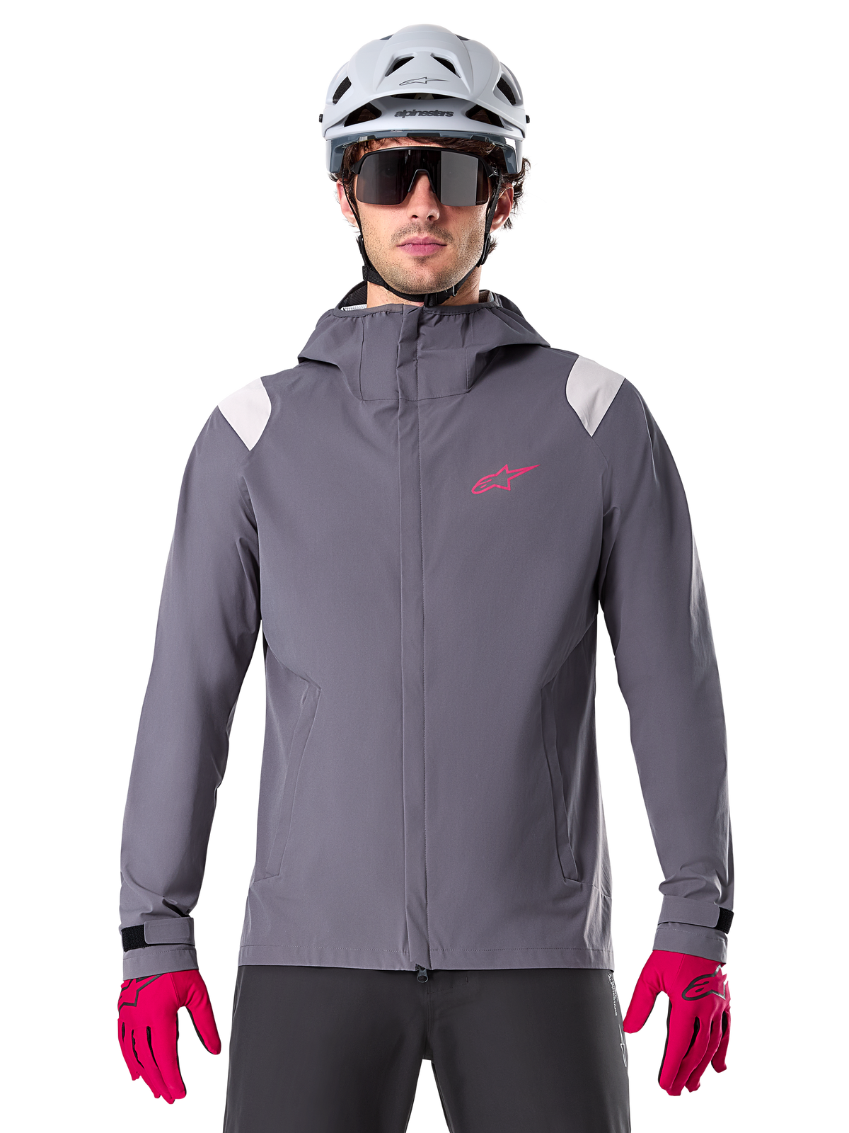 Alpinestars A-Dura Regenjacke, Mountainbike-Regenjacke, Dunkles Titanium-Grau, Kapuzendesign mit pinkem Alpinestars-Logo und hellgrauen Schultereinsätzen, getragen mit weißem MTB-Helm und leuchtend pinken Handschuhen, wasserdichte Ausrüstung für den Offroad-Radsport
