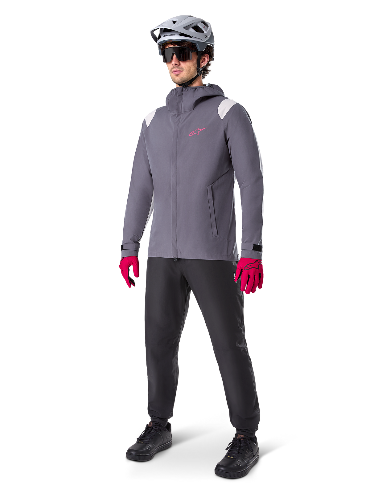 Männliches Model in der Alpinestars A-Dura Regenjacke in Dark Titanium, graue Kapuzenjacke mit pinkem Brustlogo und hellgrauen Schultereinsätzen, kombiniert mit einem grauen MTB-Helm, dunkler Sonnenbrille, leuchtend pinken Handschuhen und einer schwarzen Hose für den Offroad-Radsport.