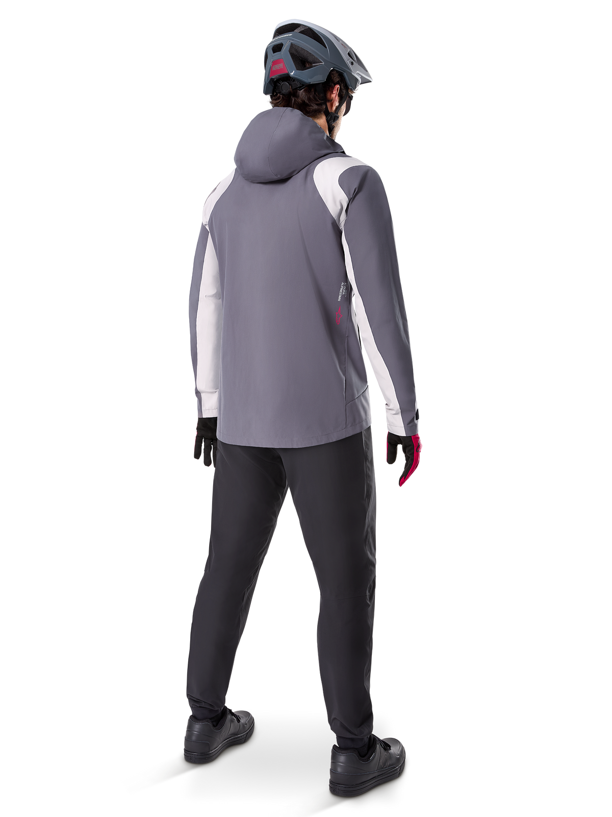 Alpinestars A-Dura Regenjacke, Mountainbike-Jacke, Dunkles Titangrau mit weißen Einsätzen, Rückansicht eines Fahrers mit passendem grauen Helm und schwarzer Hose, technische Outdoor-Ausrüstung für nasses Wetter