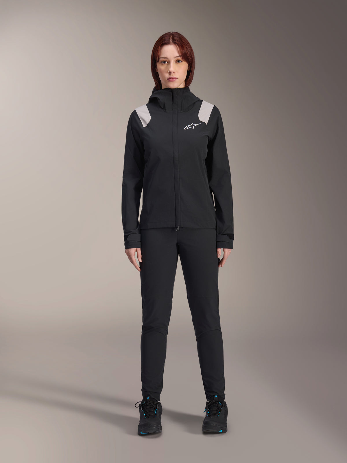 Alpinestars Stella A-Dura Regenjacke, Damen MTB-Regenjacke, Schwarz mit hellgrauen Schulterakzenten und weißem Logo, getragen mit passender schwarzer Hose und Schuhen, mit Kapuze und verstellbaren Manschetten, entwickelt für Mountainbike-Trailfahrten