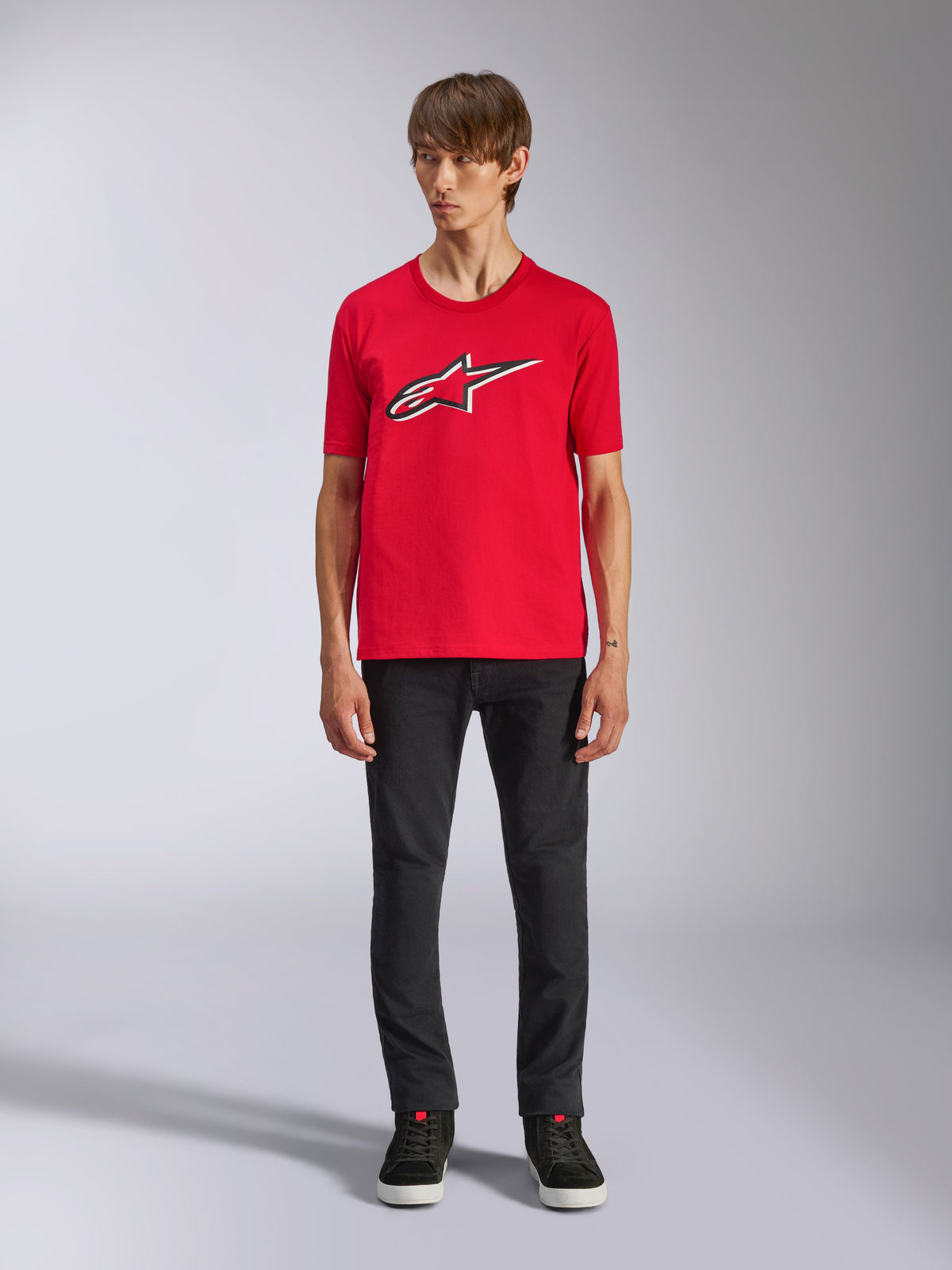 Alpinestars Ageless Shadow CSF Tee, Kurzarm-T-Shirt, Rot, vorderseitiges Ageless-Logo mit weißen und schwarzen Umrissen, getragen von einem Modell mit schwarzer Hose und Sneakern, lässige Motorsport-Streetwear