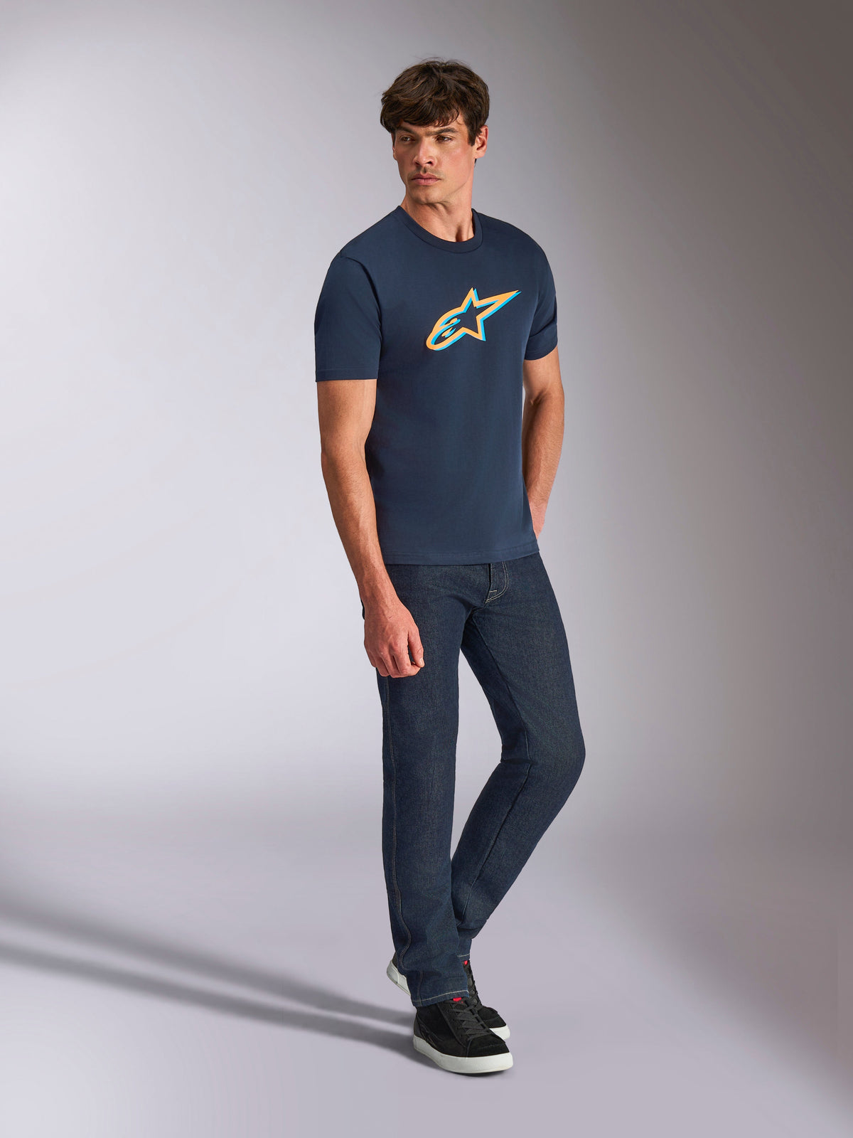 Alpinestars Ageless Shadow CSF Tee, Kurzarm-T-Shirt, Navyblau mit orange- und tealfarbenem Schattenlogo, getragen von einem Model mit dunklen Denim-Jeans und schwarzen Sneakern, lässige Motorsport-Streetwear