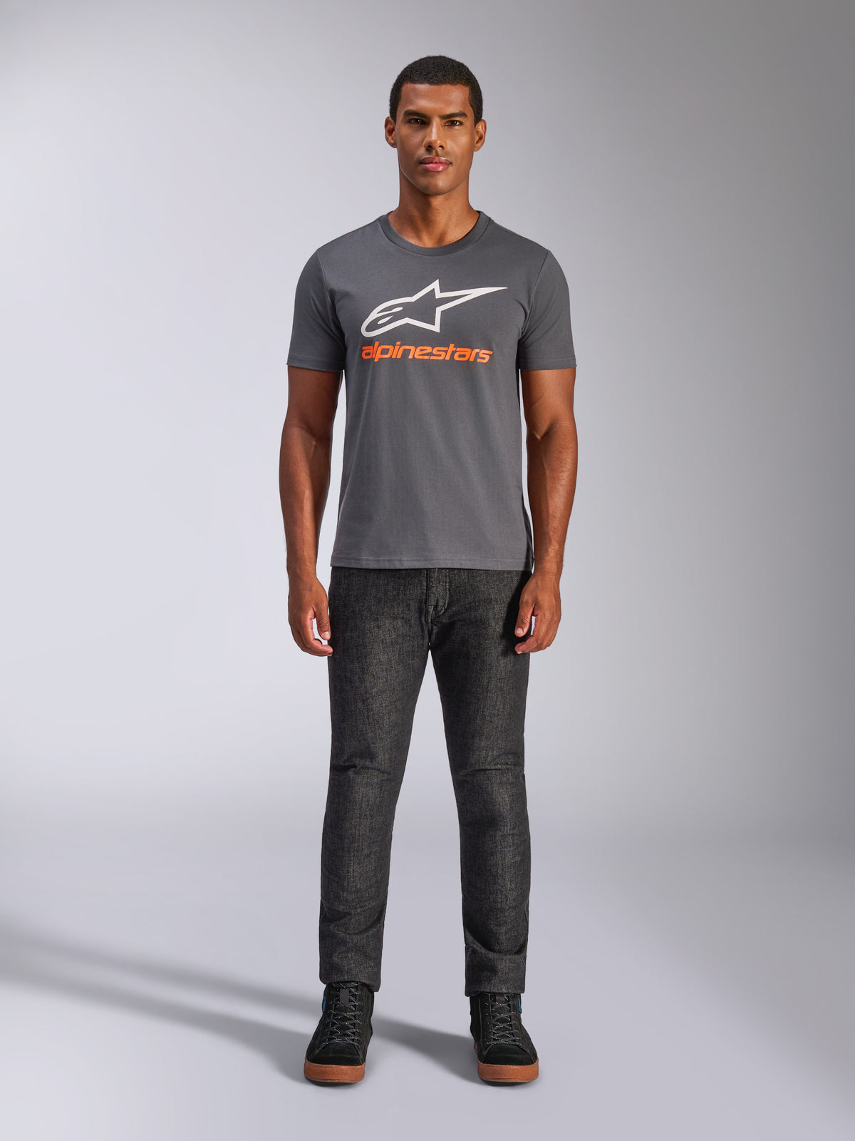 Alpinestars Always 2.0 CSF Tee, Kurzarm-T-Shirt, Anthrazitgrau mit weißem Logo und orangefarbenem Schriftzug, getragen von Model mit dunkler Jeans, lässige Motorsport-Streetwear