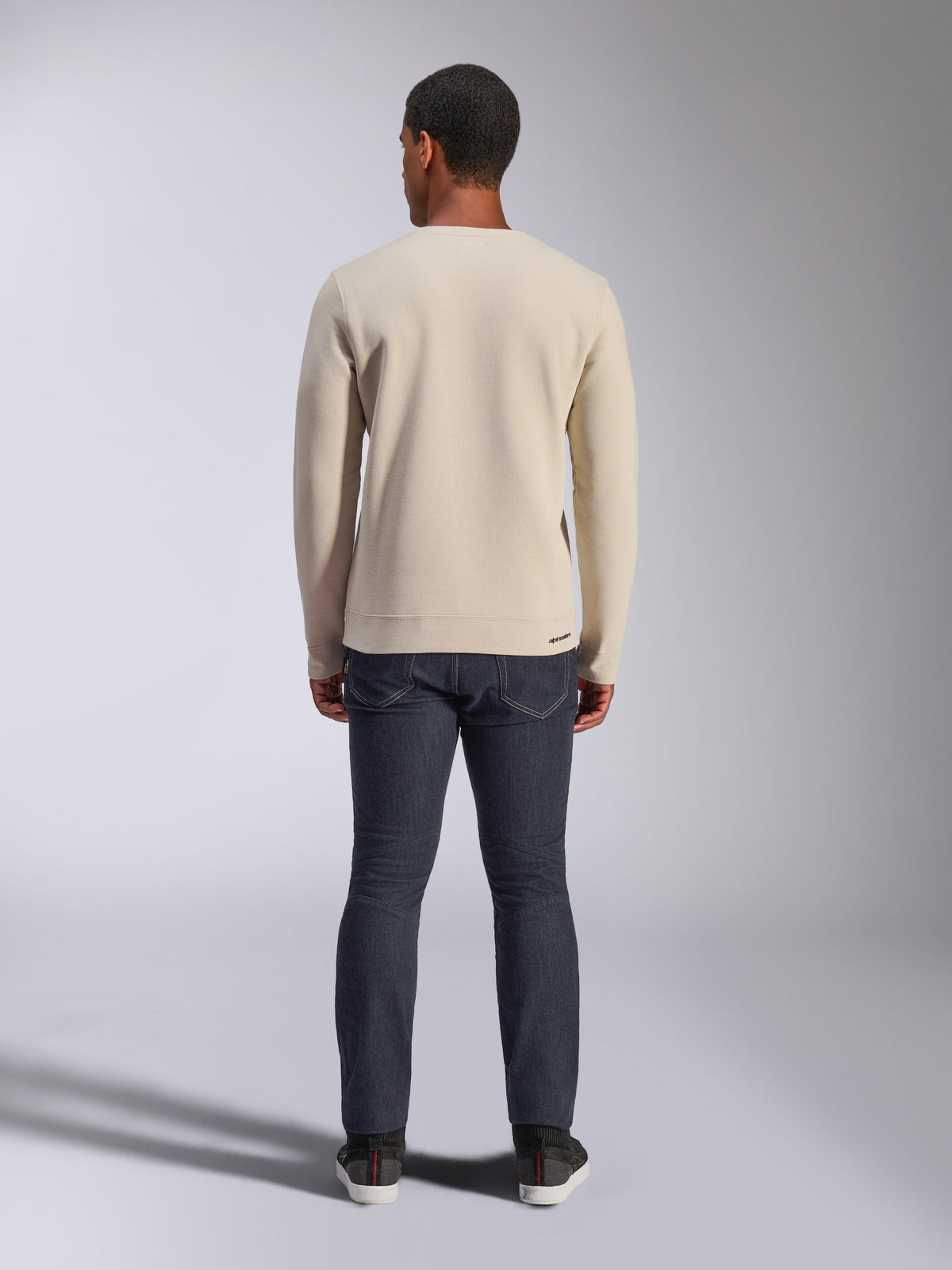 Alpinestars Positive Crew Sweatshirt, Off White, Rückansicht eines Mannes in einem beigen Rundhals-Pullover mit geripptem Saum und Bündchen, kombiniert mit dunklen Jeans, lässige motorsportinspirierte Streetwear.