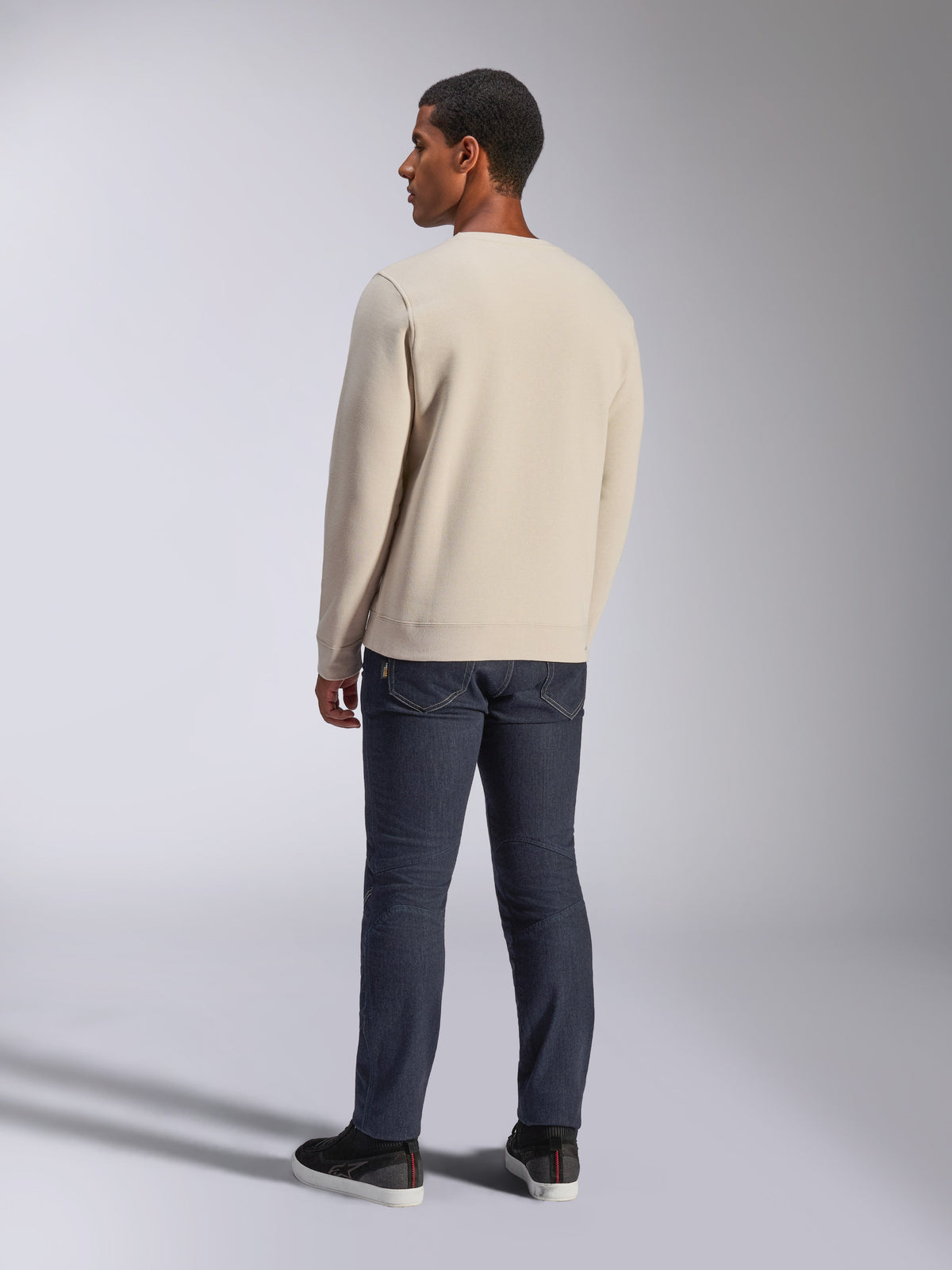 Alpinestars Positive Crew, Off-White Rundhals-Sweatshirt, Rückansicht am Modell, kombiniert mit dunklen Denim-Jeans und schwarzen Sneakern, lässige Motorsport-Streetwear, mit gerippten Bündchen und Saum