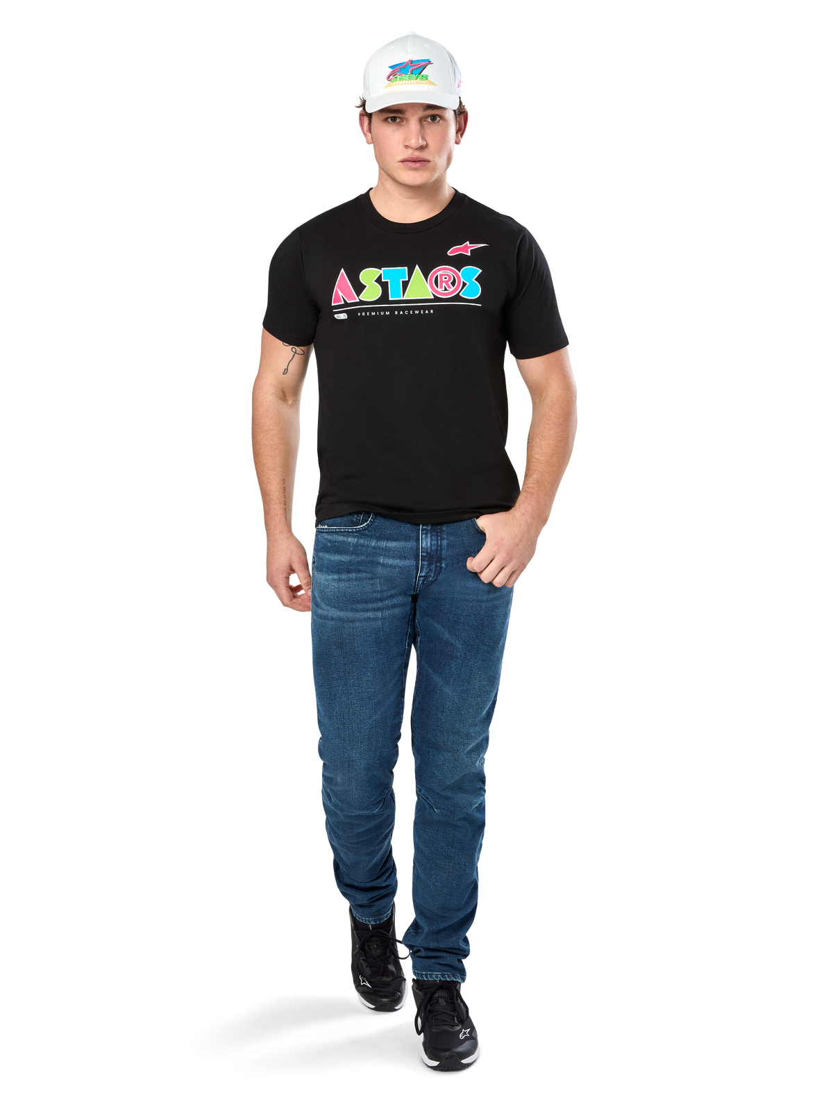 Männliches Model im Alpinestars Cereal CSF Tee, schwarzes Kurzarm-T-Shirt mit farbenfrohem geometrischem ASTARS-Logo, kombiniert mit blauen Jeans und weißer Kappe, lässige motorsport-inspirierte Streetwear.