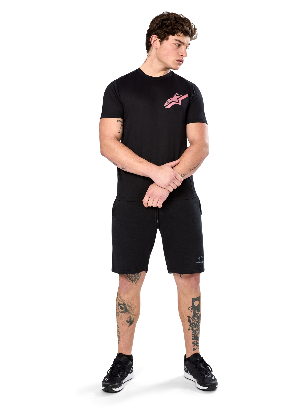 Model trägt Alpinestars Attrition Performance Tee, kurzärmliges Performance-T-Shirt, Schwarz mit pinkem Astars-Logo auf der Brust, kombiniert mit schwarzen Shorts und Sneakern, lässige motorsport-inspirierte Sportbekleidung