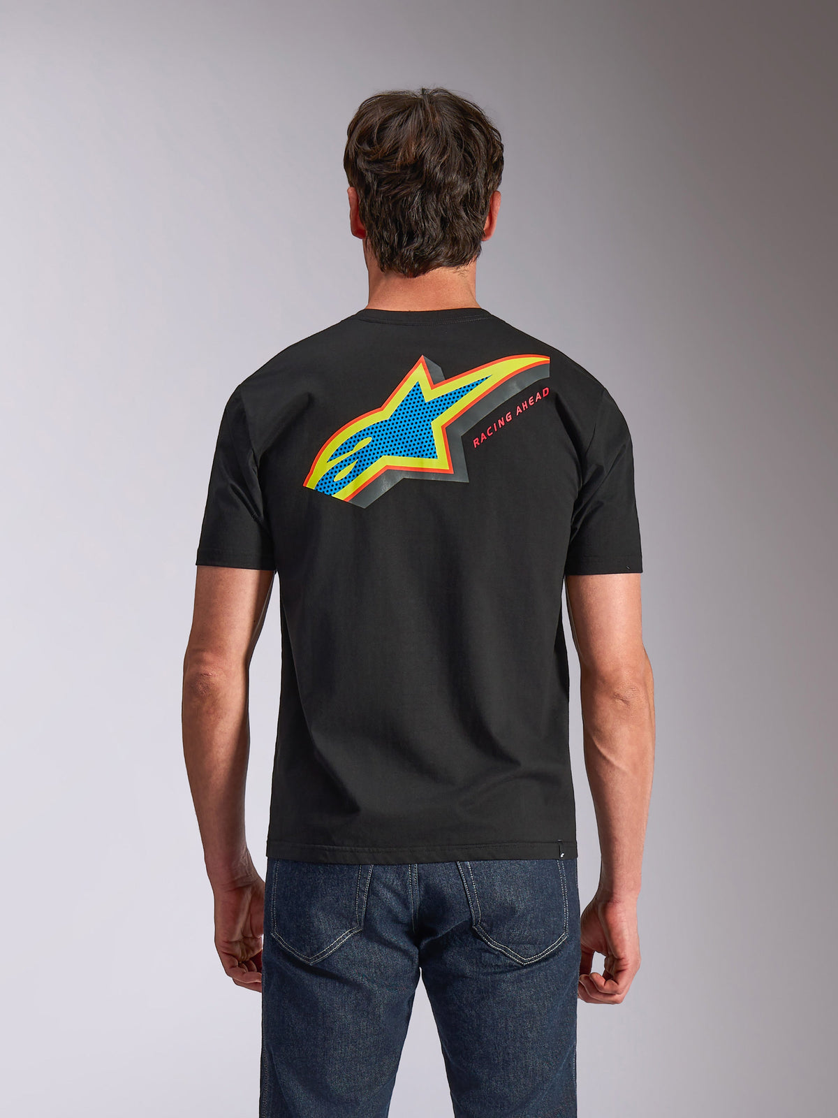 Alpinestars Angler CSF Tee, kurzärmeliges T-Shirt, Schwarz, Rückansicht mit lebendigem mehrfarbigem Astars-Logo mit perforierter Füllung und „RACING AHEAD“-Schriftzug, getragen mit blauen Jeans, lässige Motorsport-inspirierte Bekleidung.