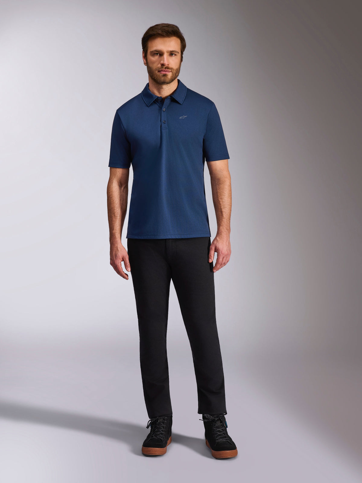 Alpinestars Impetus Sport Polo, Kurzarm-Poloshirt, Marineblau, kleines silbernes Astars-Logo auf der Brust, Drei-Knopf-Leiste, getragen von einem Modell mit schwarzer Hose und Stiefeln, lässige vom Motorsport inspirierte Sportbekleidung