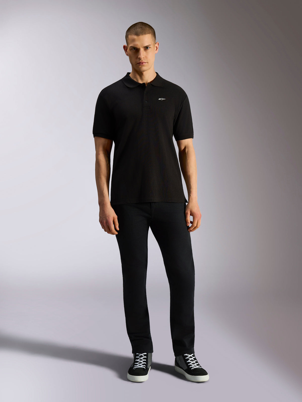Alpinestars Endeavor Classic Polo, kurzärmeliges Piqué-Poloshirt, Schwarz, kleines weißes Astars-Logo auf der Brust, Rippkragen und Ärmelbündchen, getragen von einem Model mit schwarzer Hose und Sneakern, lässige Motorsport-Streetwear
