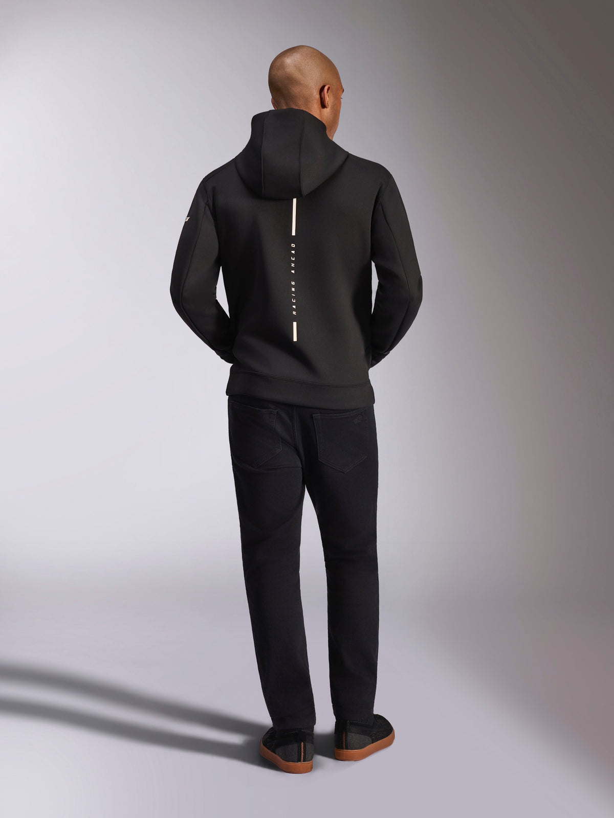 Alpinestars Insight Kapuzenpullover, Schwarz, Rückansicht am Modell, mit einem vertikalen weißen Streifen-Logo auf dem Rücken und dem Schriftzug „Racing Ahead“, legerer Sportswear-Hoodie aus Tri-Blend-Doppelvlies, kombiniert mit einer schwarzen Hose.
