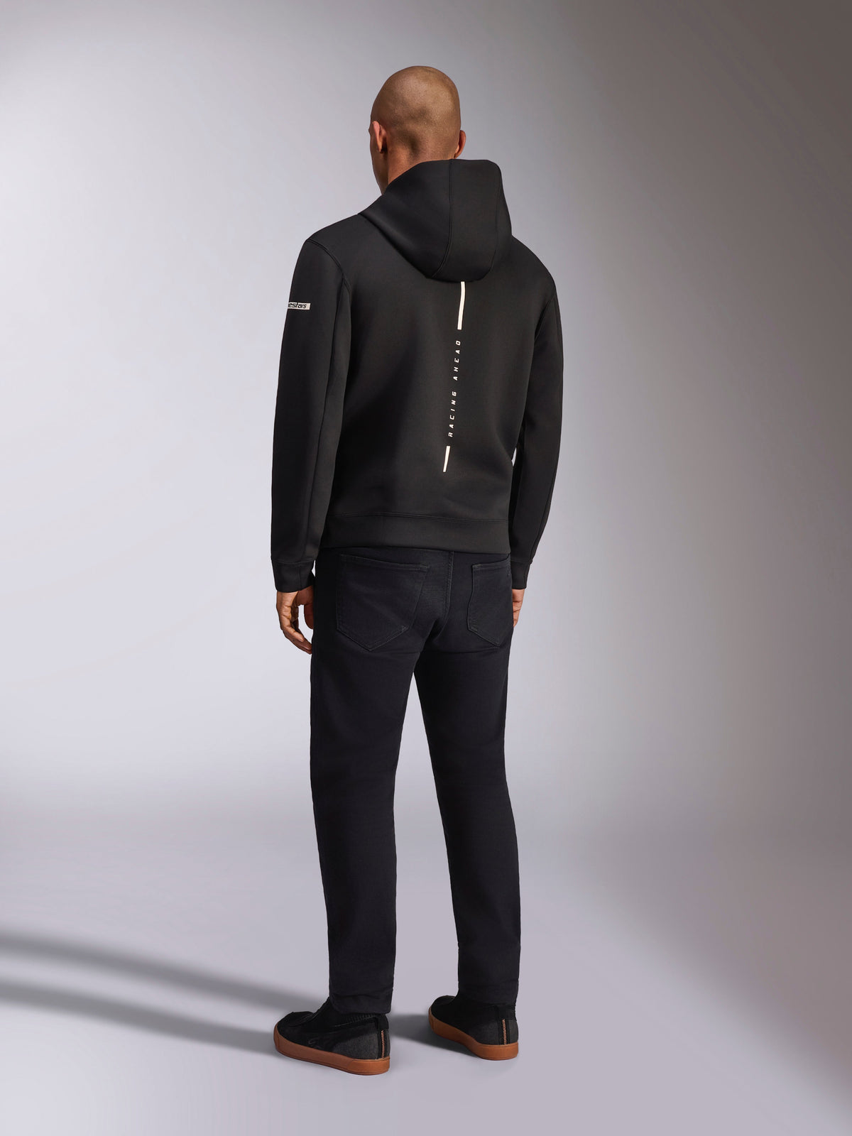 Alpinestars Insight Hoodie, schwarzer Sport-Kapuzenpullover, Rückansicht mit vertikalem weißem Streifen und „RACING AHEAD“-Schriftzug, Ärmellogo, kombiniert mit schwarzer Hose und Sneakern, lässiges, vom Motorsport inspiriertes Design