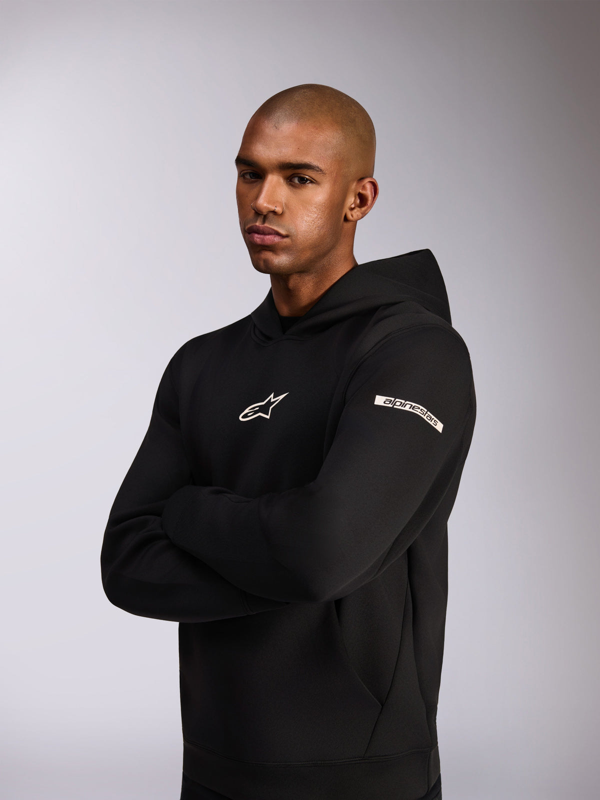 Alpinestars Insight Hoodie, lässiger Sportswear-Kapuzenpullover, Schwarz, weißes Astars-Logo auf der Brust und Wortlogo am Oberärmel, Model trägt Kapuzenpullover mit verschränkten Armen, hochwertige Doppelvlies-Konstruktion