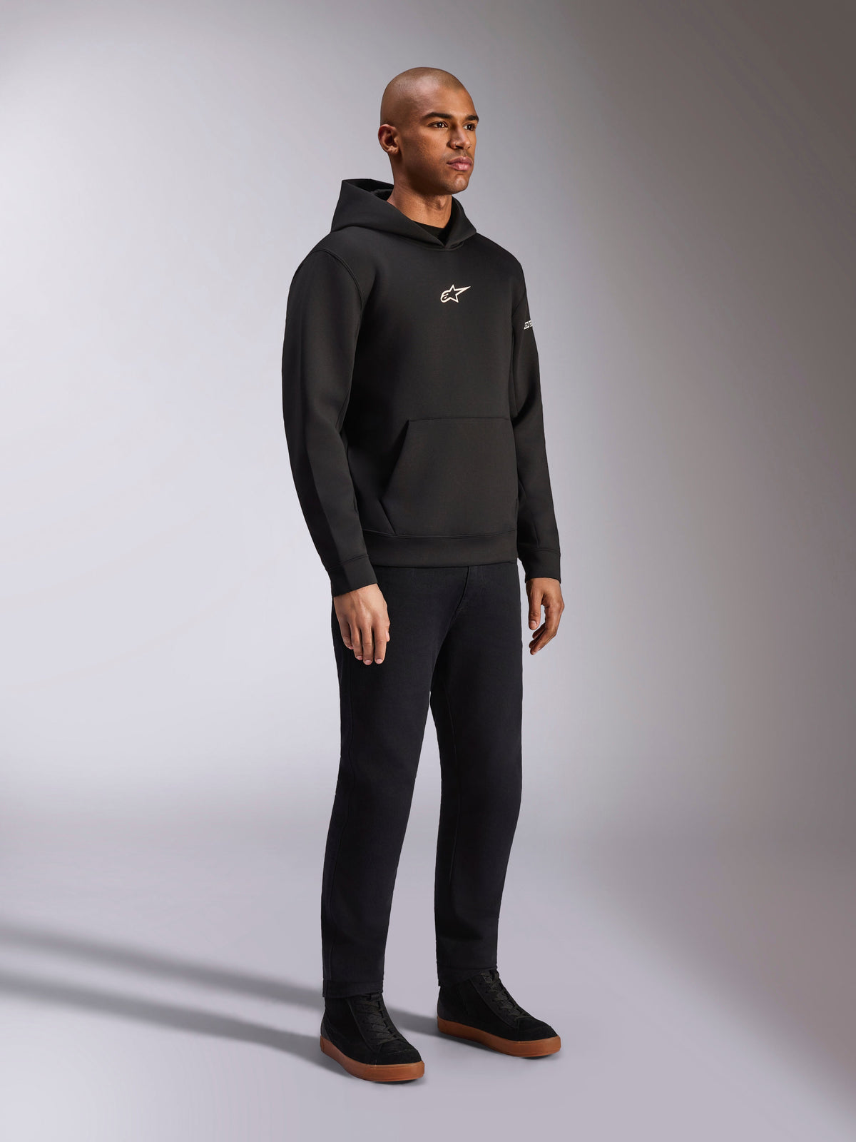 Alpinestars Insight Hoodie, Schwarz, lässiger Fleece-Kapuzenpullover mit weißem Astars-Logo auf der Brust, Kängurutasche, getragen mit schwarzer Hose und Sneakern, Ganzkörper-Studioaufnahme, motorsport-inspirierte Streetwear