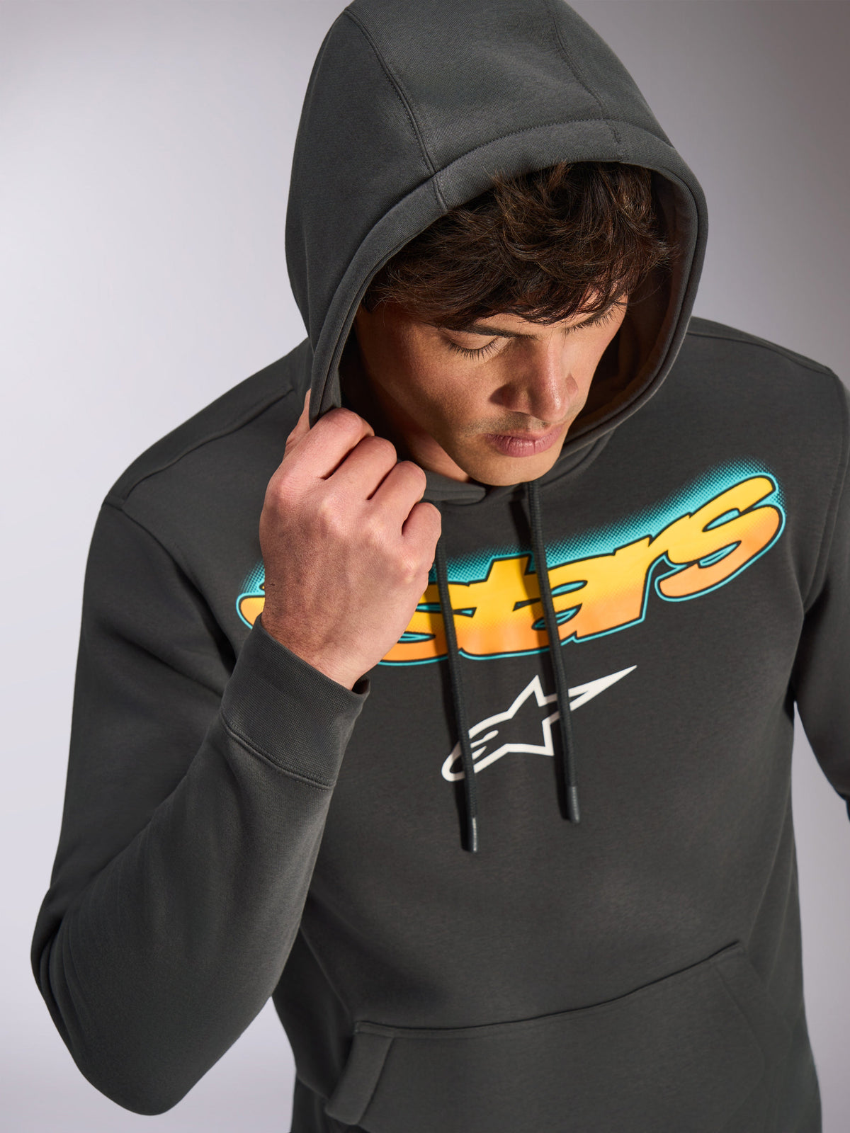 Alpinestars Ellipsoid Kapuzenpullover, Gunmetal-Grau, lässiger Kapuzenpullover mit einem Astars-Schriftzug im Farbverlauf von Cyan nach Orange und weißem Logo, getragen von einem Model mit aufgesetzter Kapuze, Motorsport-inspirierte Streetwear