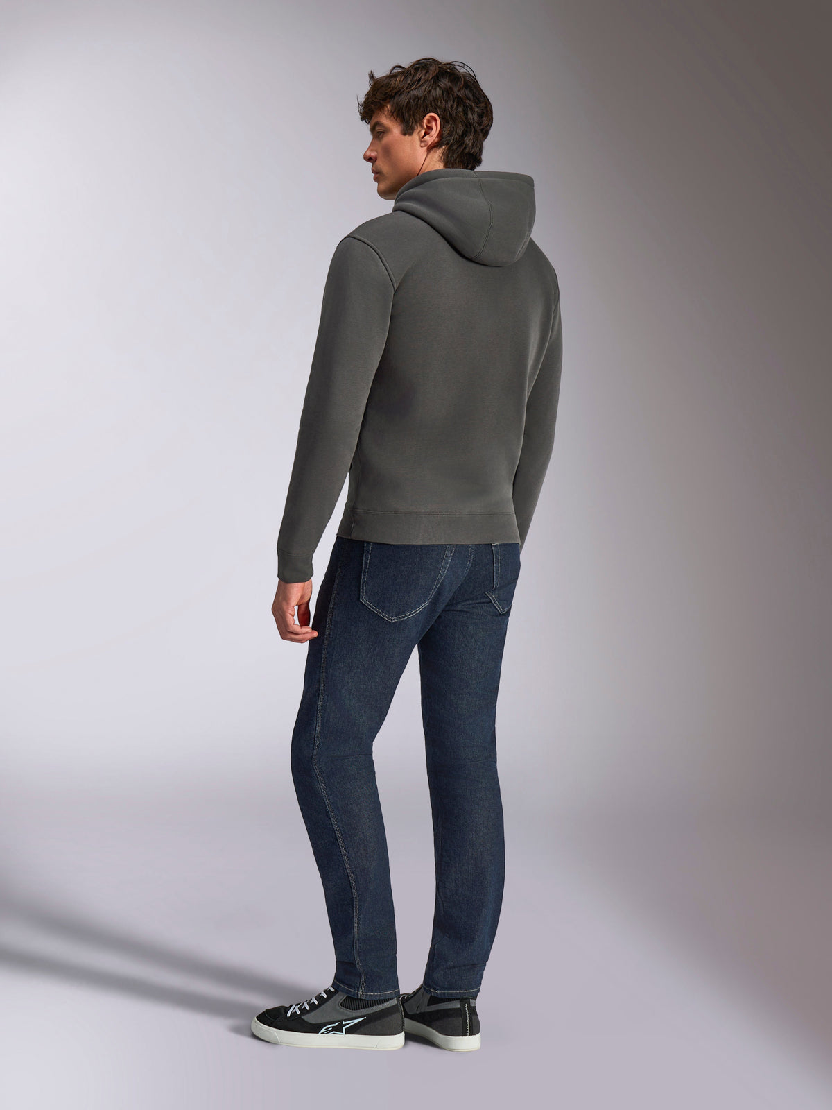 Alpinestars Ellipsoid Hoodie, lässige Sportbekleidung, Gunmetal-Grau, Rückansicht an männlichem Model, Kapuze mit Kordelzug, lange Ärmel, Rippbündchen an Ärmeln und Saum, kombiniert mit dunkler Jeans und Sneakern