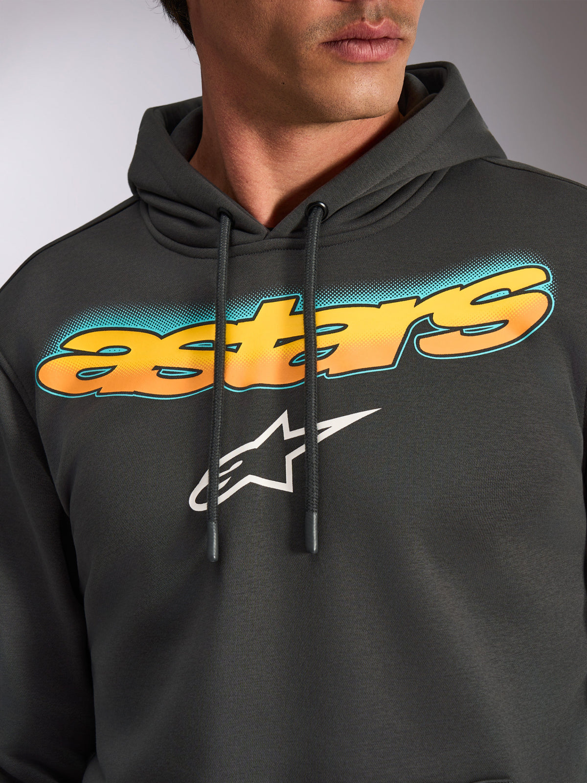Alpinestars Ellipsoid Hoodie, Gunmetal-Grau, Nahaufnahme der Brust mit geschwungenem „astars“-Schriftzug, Gelb-Orange-Farbverlauf und blaugrünen Akzenten, weißes Alpinestars Stern-Logo, lässiges, vom Motorsport inspiriertes Streetwear-Fleece