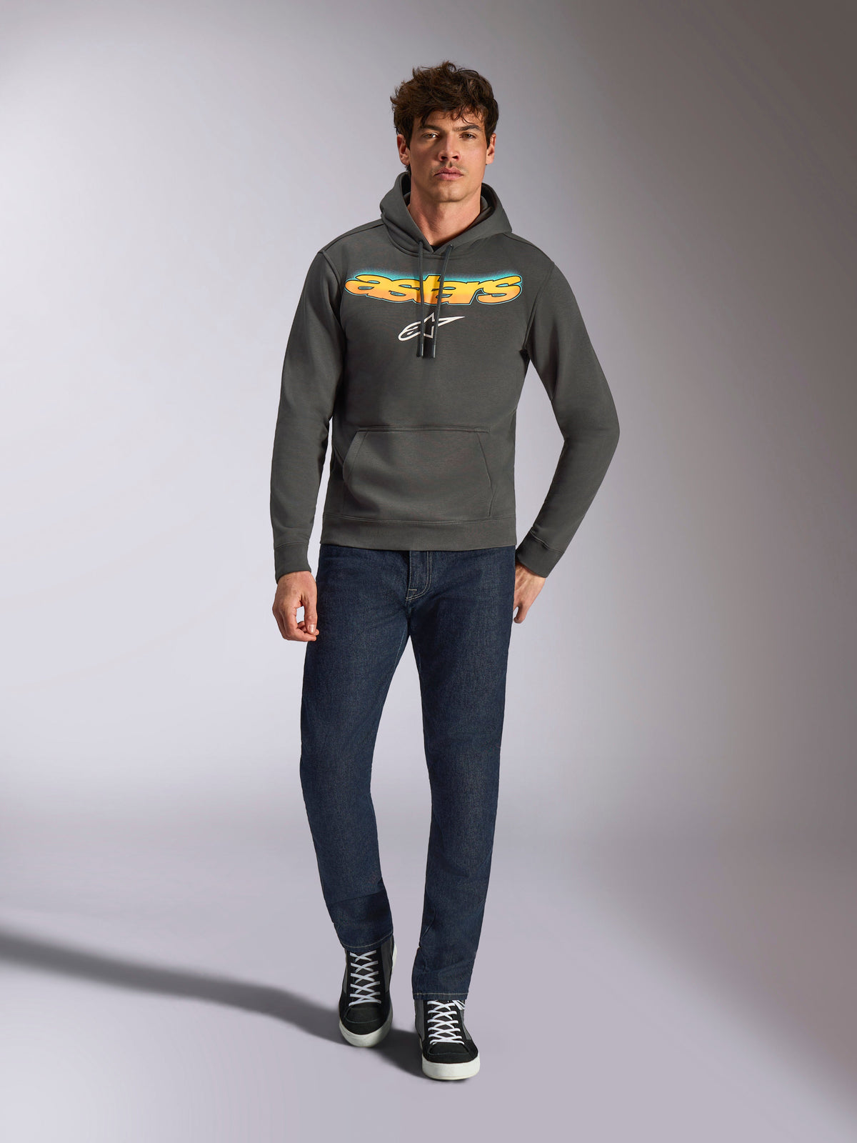 Alpinestars Ellipsoid Hoodie, lässige Sportbekleidung, gunmetal-grau mit orange-gelbem Astars-Logo mit Farbverlauf und weißem Symbol, Kapuze mit Kordelzug, getragen von einem männlichen Model mit dunkelblauen Jeans und schwarzen Sneakern, moderne, vom Motorsport inspirierte Streetwear