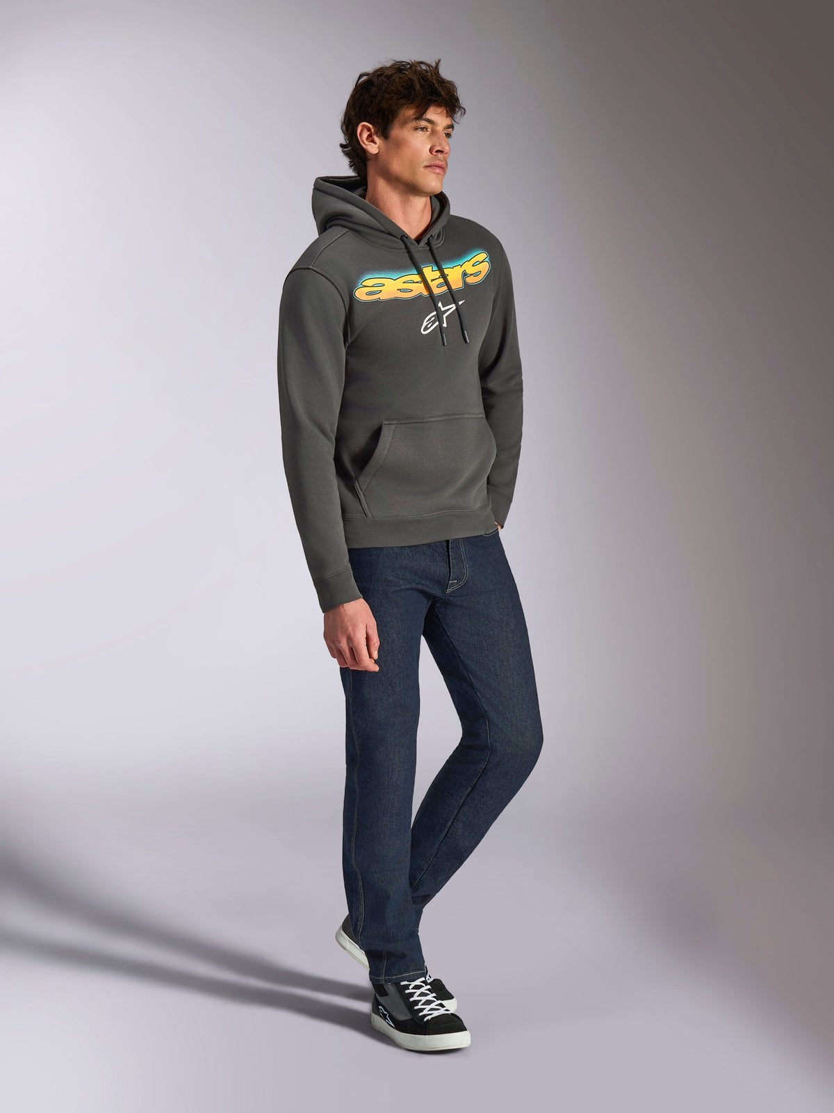 Alpinestars Ellipsoid Hoodie, Fleece-Kapuzenpullover in Gunmetal-Grau, vorne gebogener Astars-Schriftzug mit blau-gelbem Farbverlauf und weißem Logo, kombiniert mit dunkler Jeans und schwarzen Sneakern, lässige Streetwear.
