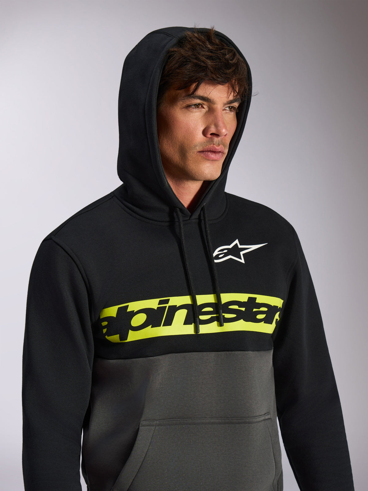 Alpinestars Parallel Hoodie, lässige Streetwear im Color-Block-Design, oben Schwarz und unten Gunmetal-Grau, neongelber Bruststreifen mit schwarzem Alpinestars-Wortlogo, weißes Astars-Logo, Kapuzenpullover mit Kordelzug