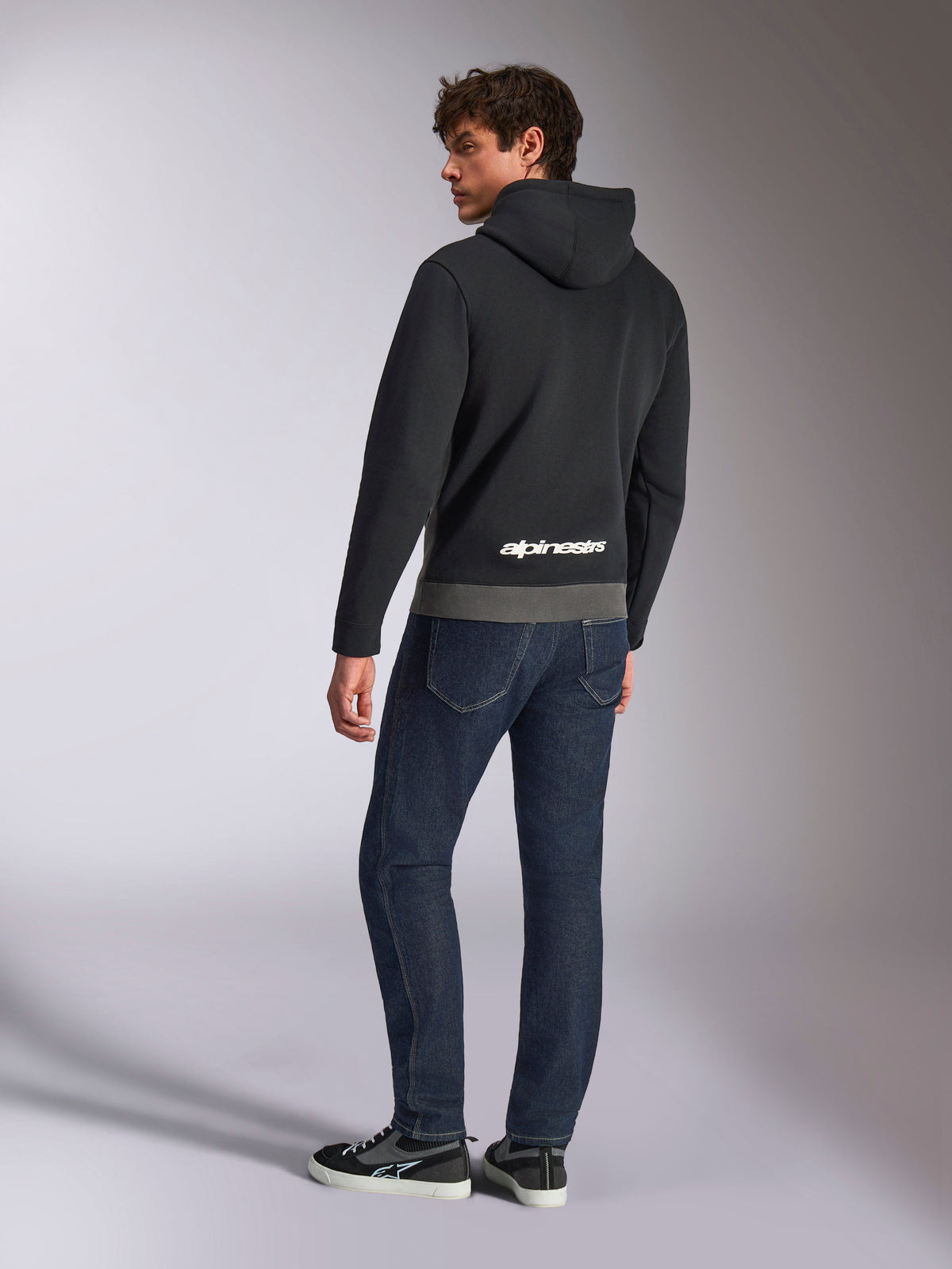 Alpinestars Parallel Hoodie, Rückansicht eines Models im schwarz- und gunmetal-grauen Kapuzenpullover, mit weißem Alpinestars-Logo auf dem unteren Rücken, kombiniert mit dunkelblauen Jeans, lässige, vom Motorsport inspirierte Streetwear