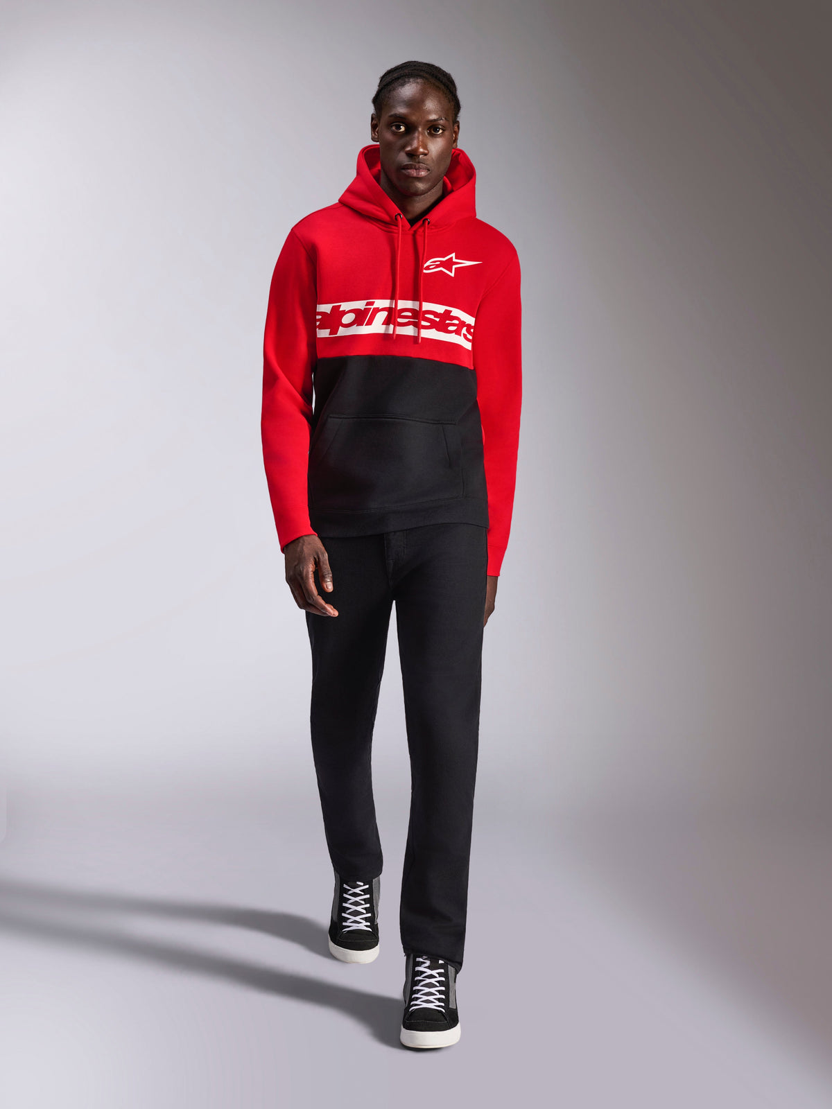Alpinestars Parallel Hoodie, color-blocked pullover fleece, Red and Black, featuring white Alpinestars word logo across the chest and Astars logo on the left chest, worn by a model with black pants and high-top sneakers, casual motorsport streetwear