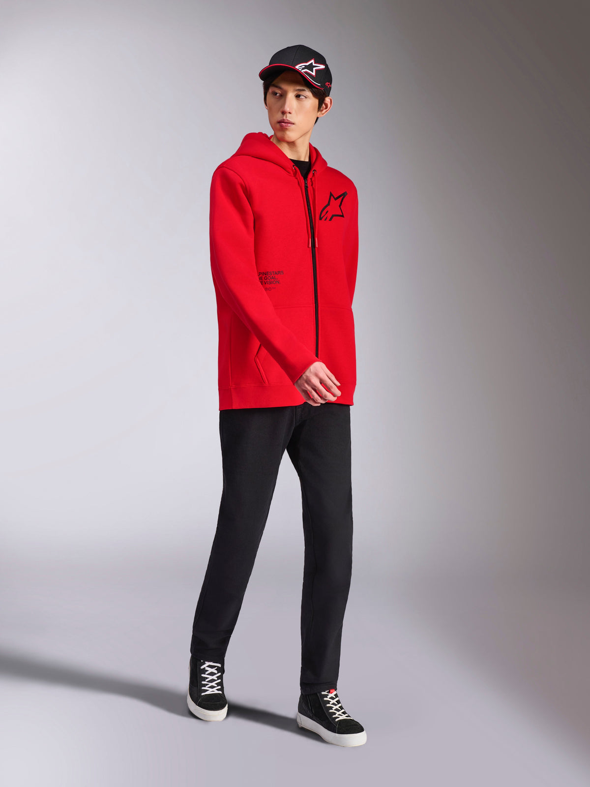 Alpinestars Transverse Hoodie in Red, male model wearing full-zip hooded fleece, black Astars logo and text print, styled with black pants and cap, casual motorsport streetwear