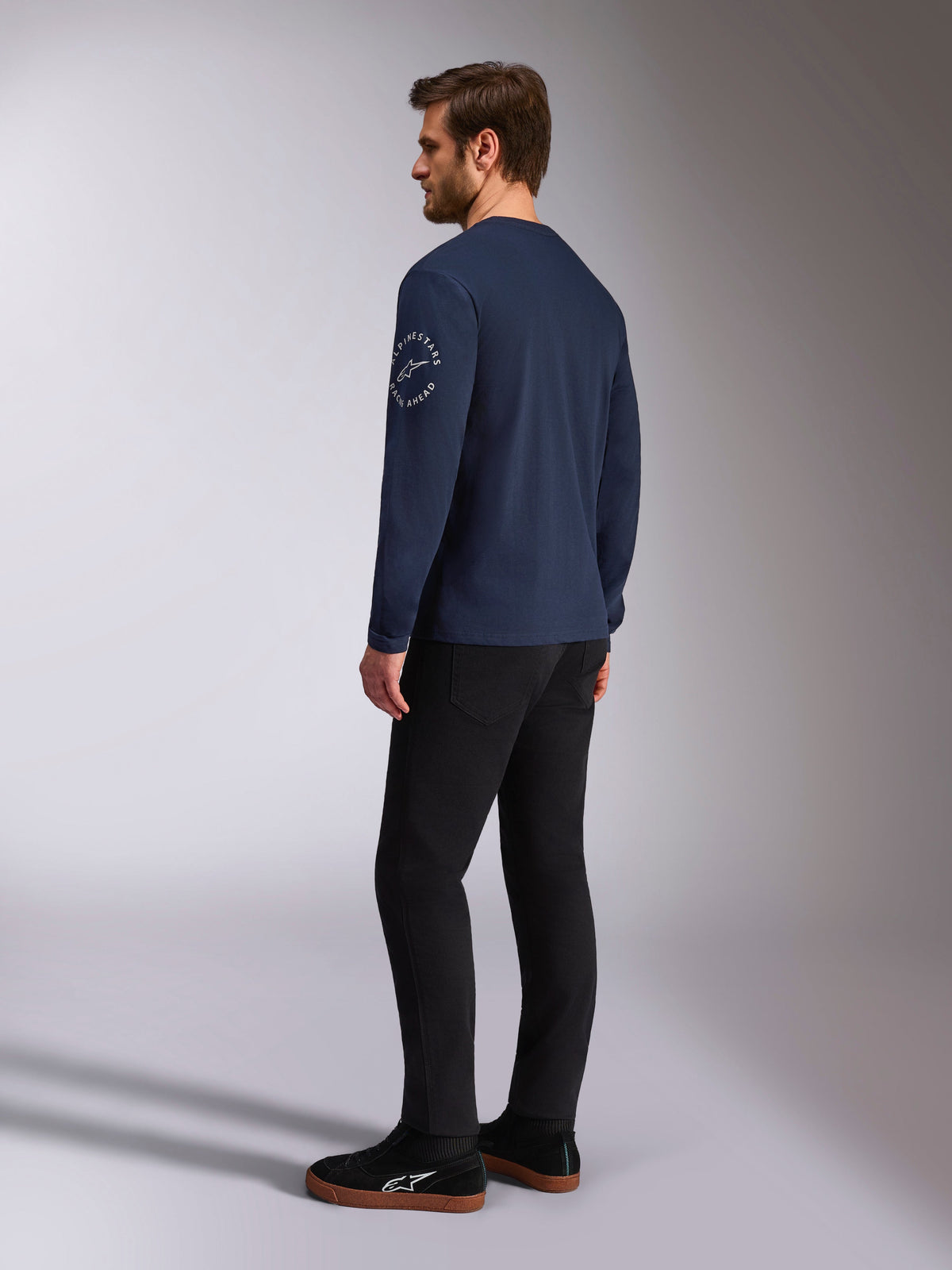 Alpinestars Mandatory CSF Langarmshirt, Navy, weißes kreisförmiges Logo am Ärmel, Rückansicht, getragen mit schwarzer Hose und schwarzen Sneakern, lässige Motorsport-Streetwear