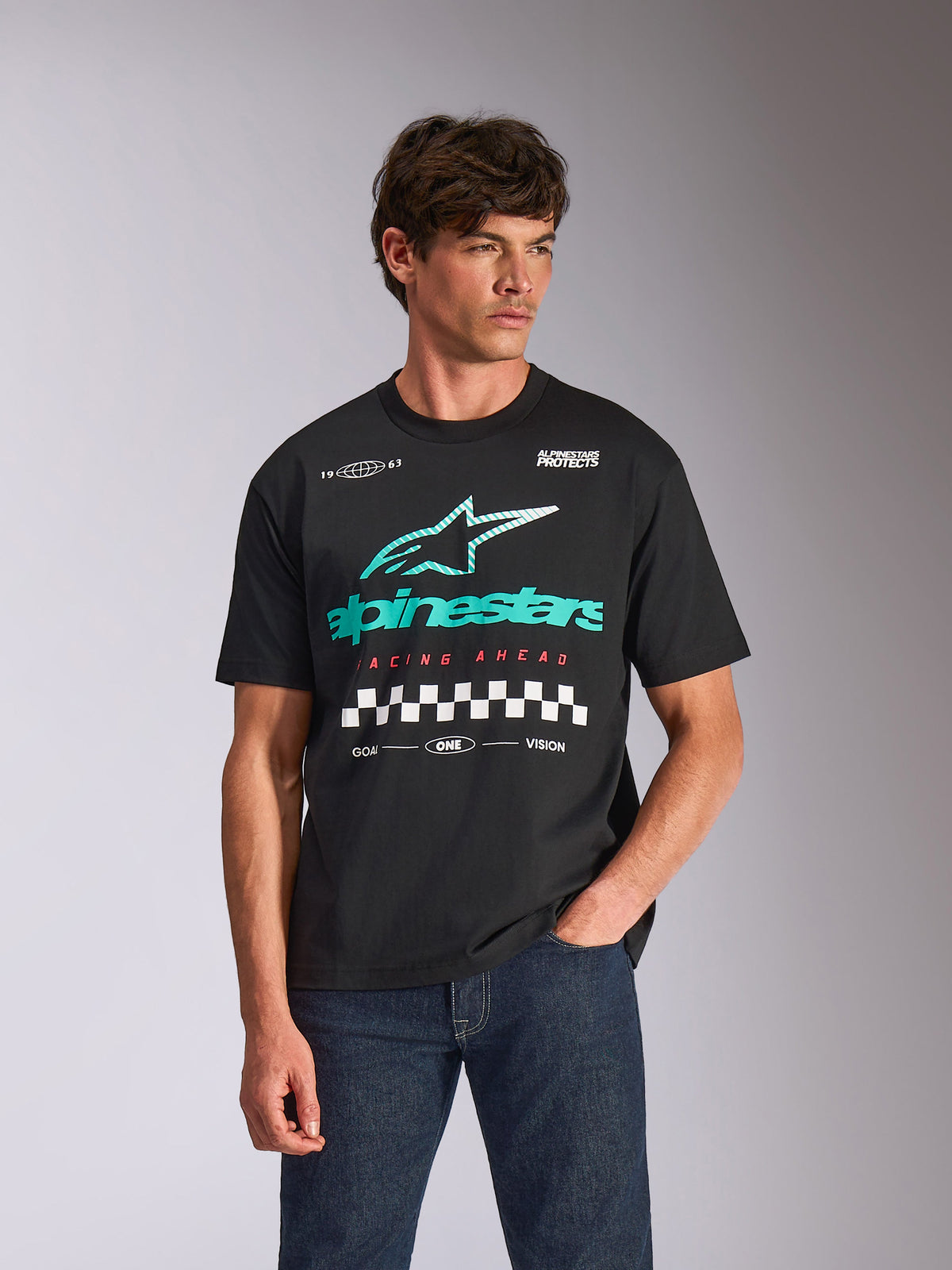 Alpinestars Aplomb Oversized CSF Tee, Kurzarm-T-Shirt, Schwarz, mit einem großen blaugrünen und weißen Astars-Kombi-Logo und einer Schachbrett-Grafik, getragen von einem männlichen Model mit dunkelblauen Jeans, lässige Motorsport-Streetwear