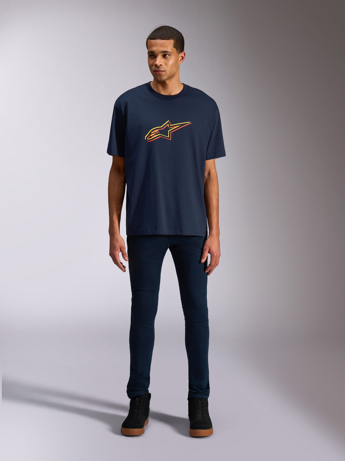 Alpinestars Alter Oversized CSF Tee, Kurzarm-T-Shirt, Marineblau, mit einem Alpinestars-Sternlogo auf der Vorderseite mit gelbem und orangem Farbversatz, getragen von einem Model mit dunkler Jeans und schwarzen Sneakern, lässige Lifestyle-Sportbekleidung