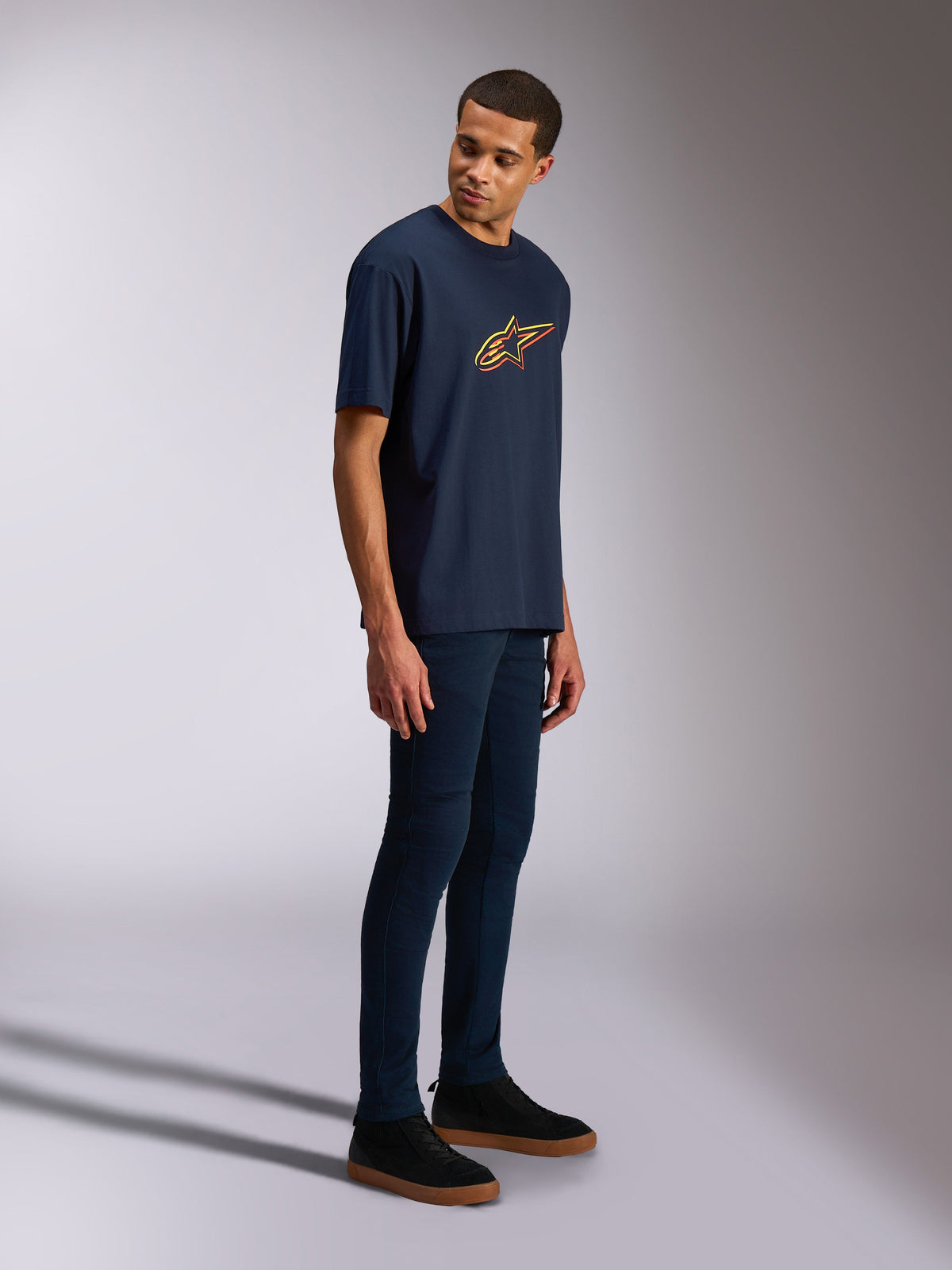 Alpinestars Alter Oversized CSF Tee, kurzärmeliges T-Shirt, Marineblau, gelbes und oranges Astars-Logo, getragen von einem Model mit dunklen Jeans und schwarzen Sneakern, lässige motorsport-inspirierte Streetwear