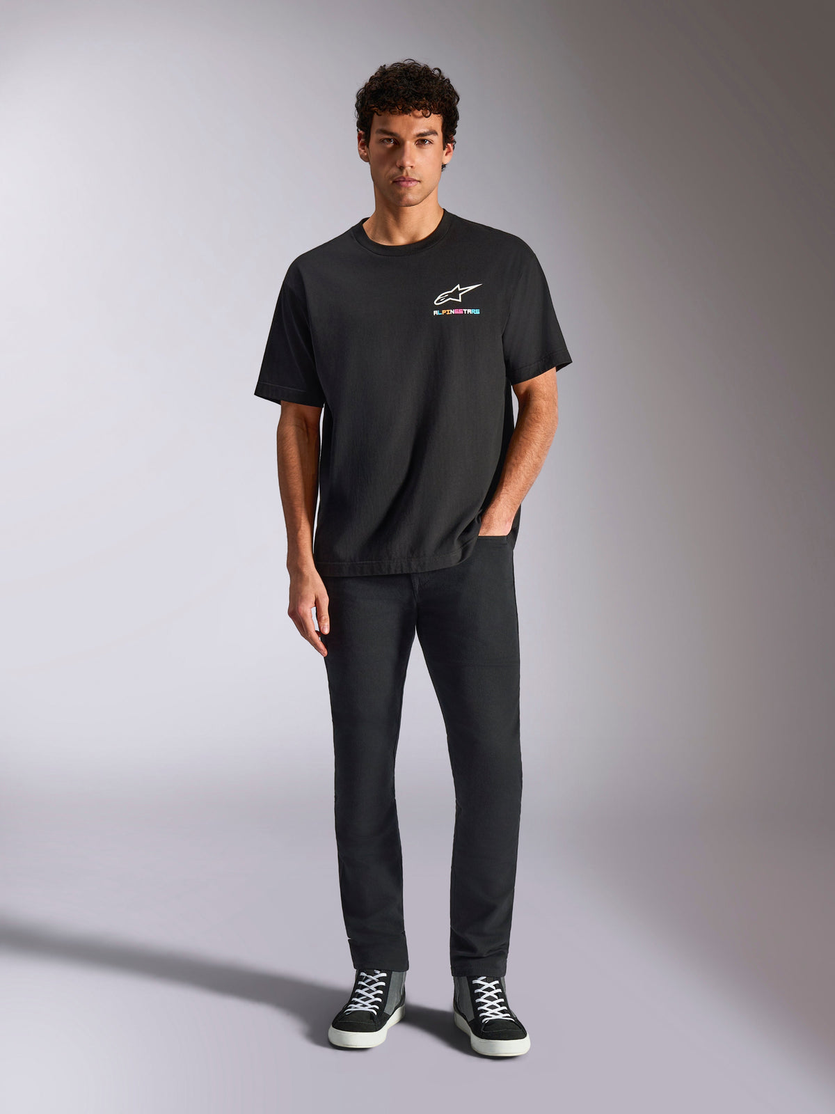 Alpinestars Wild Oversized CSF Tee, kurzärmeliges T-Shirt, Schwarz, getragen von einem Model mit schwarzer Hose und Sneakern, mit weißem Astars-Logo und lebhaftem, mehrfarbigem Branding, lässige Motorsport-Streetwear
