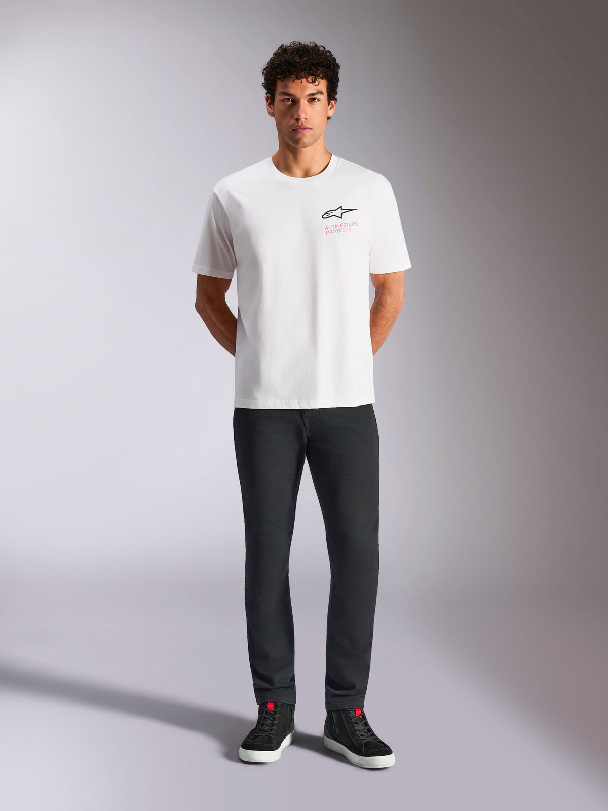 Alpinestars Pop Helmet CSF Tee, Short Sleeve T-Shirt, White, worn by a model with black pants and black high-top sneakers, front Astars logo with pink 'Alpinestars Protects' text, casual motorsport streetwear