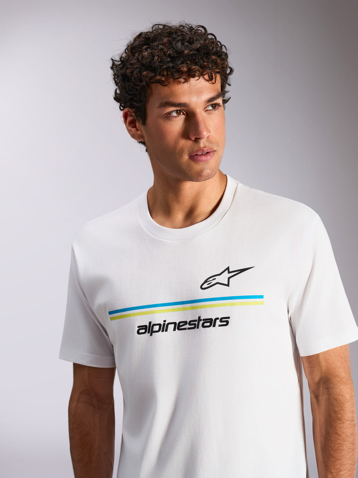 Alpinestars Lanes CSF Tee, kurzärmeliges T-Shirt, Weiß, mit blauen und limettengrünen horizontalen Streifen sowie schwarzem Astars-Logo und Schriftzug, getragen von einem Model mit lockigem Haar, lässige vom Motorsport inspirierte Streetwear