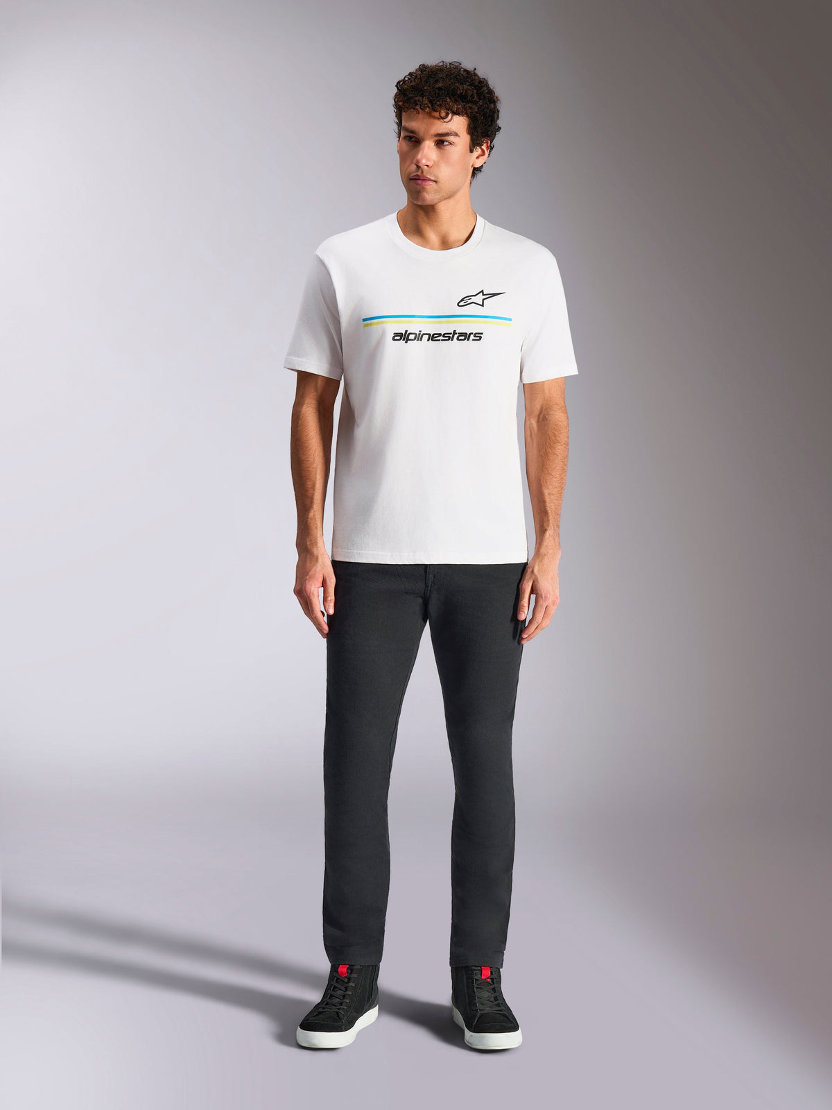 Alpinestars Lanes CSF Tee, Kurzarm-T-Shirt, Weiß, blau-gelbe Streifen-Logo-Grafik, getragen von Model mit schwarzer Hose und Sneakern, lässige Motorsport-Streetwear