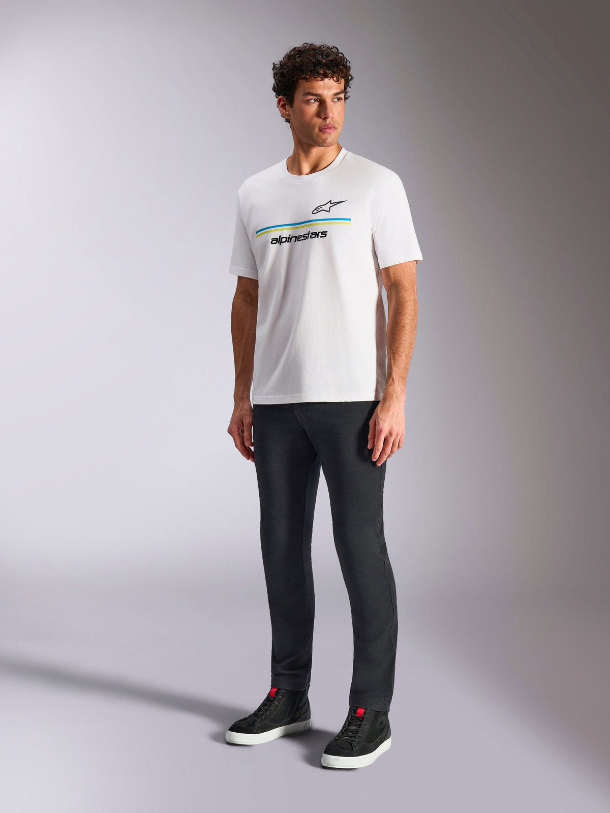 Alpinestars Lanes CSF T-Shirt, weißes Kurzarm-T-Shirt, getragen von einem Model mit schwarzer Slim-Fit-Hose und schwarzen High-Top-Sneakern, blaue und gelbe horizontale Streifen mit Alpinestars-Branding auf der Brust, lässige Motorsport-inspirierte Streetwear