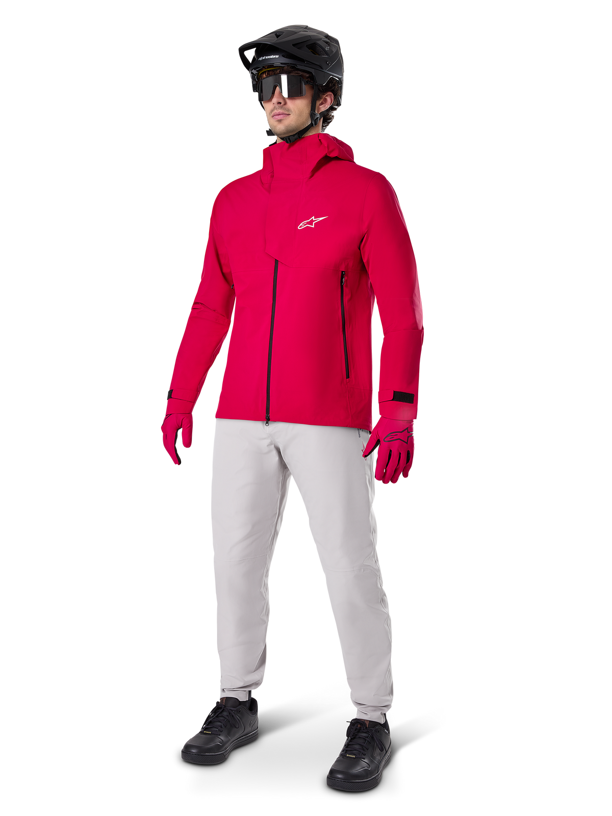 Männlicher Radfahrer in der Alpinestars A-Dura Elite wasserdichten Jacke in Red Fluo, kombiniert mit hellgrauer technischer Hose, roten Handschuhen, schwarzem MTB-Helm mit Brille und schwarzen Schuhen, komplettes Hochleistungs-Outfit für Mountainbiking