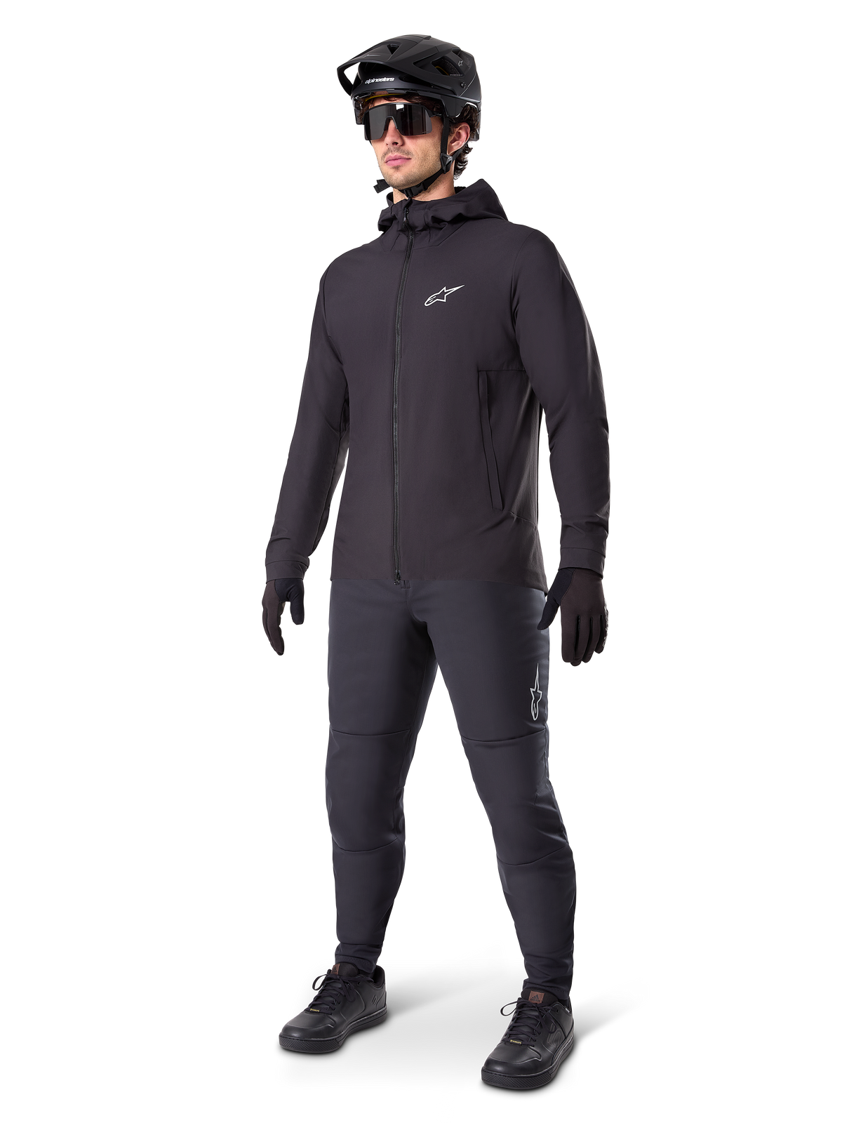 Alpinestars A-Dura Thermojacke in Schwarz, getragen von einem männlichen Fahrer mit kompletter Mountainbike-Ausrüstung inklusive schwarzem Helm, Sonnenbrille und Radhose, leichte isolierte Kapuzenjacke für Radsport-Performance bei kaltem Wetter.