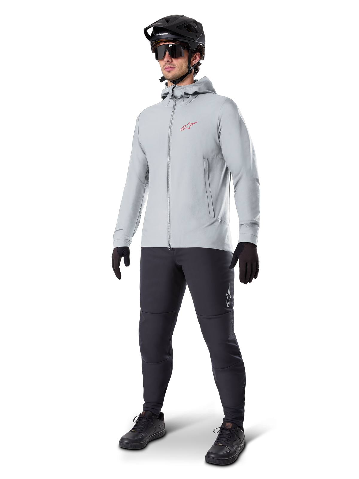 Alpinestars A-Dura Thermojacke in Titanium, hellgraue Fahrradjacke mit Kapuze, rotes Alpinestars-Logo auf der Brust, getragen von einem Mann mit schwarzem MTB-Helm, Sonnenbrille, dunkelgrauer Hose und schwarzen Handschuhen, professionelle Mountainbike-Ausrüstung