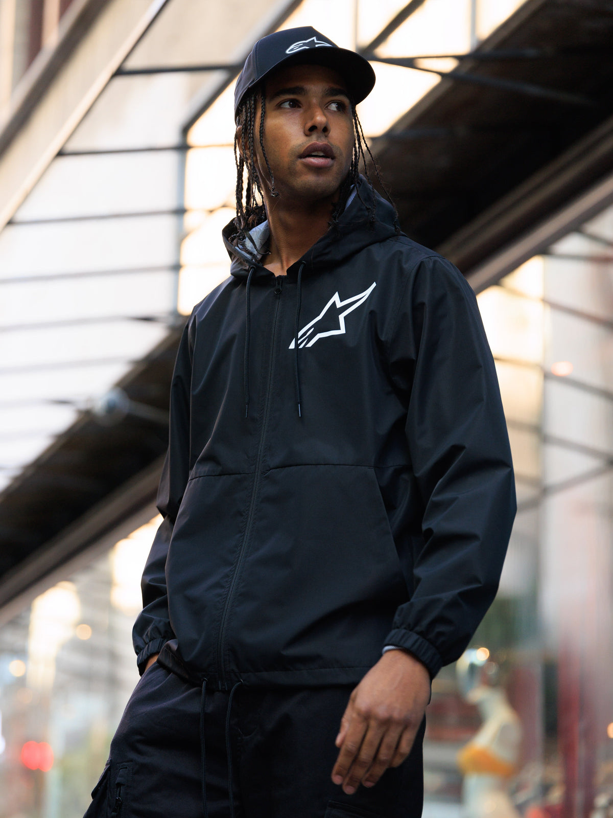 Alpinestars Speeder Chromium Windbreaker-Jacke, Schwarz, Windjacke mit Kapuze und weißem Logo auf der Brust, getragen von einem Mann mit passender schwarzer Alpinestars-Cap in einer urbanen Umgebung, lässige Motorsport-Streetwear.