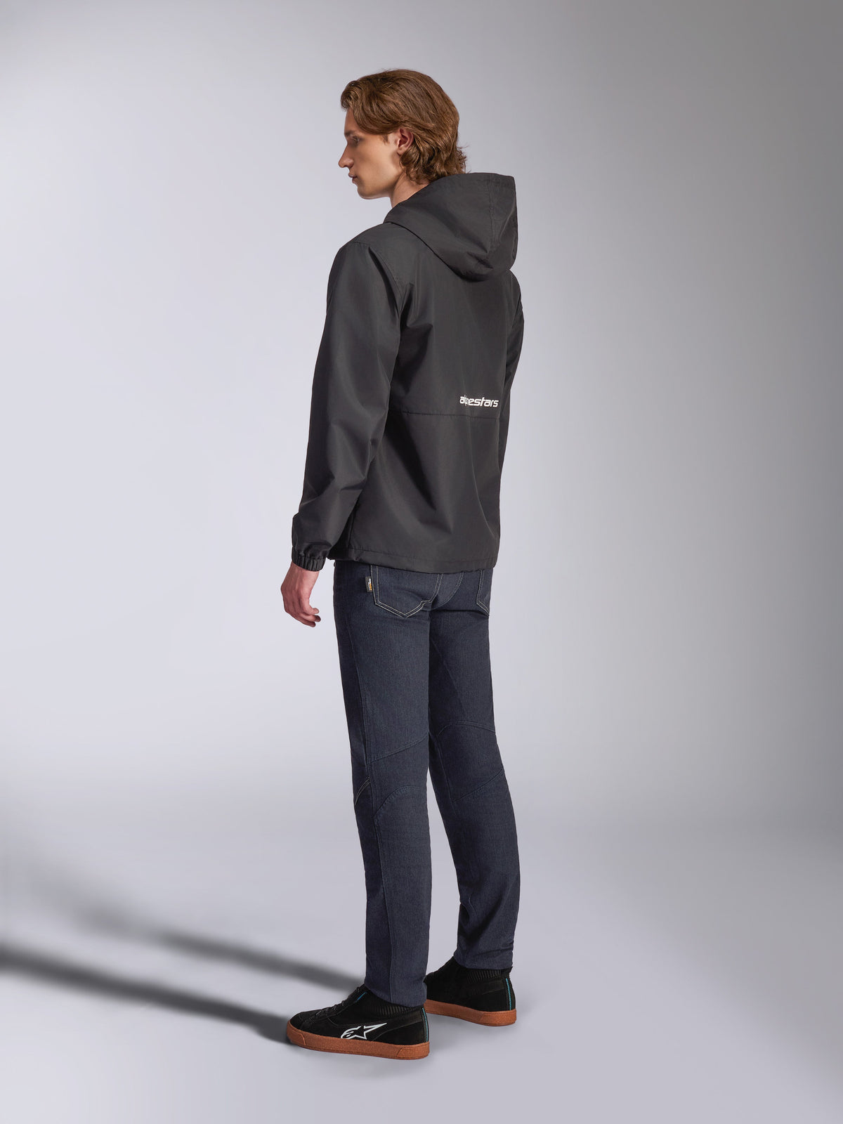 Alpinestars Speeder Chromium Windbreaker-Jacke, Schwarz mit weißem Logo auf der Rückseite, Herren-Freizeit-Windjacke mit Kapuze, Seitenansicht getragen mit dunkler Jeans und schwarzen Sneakern, Motorsport-inspirierte Streetwear