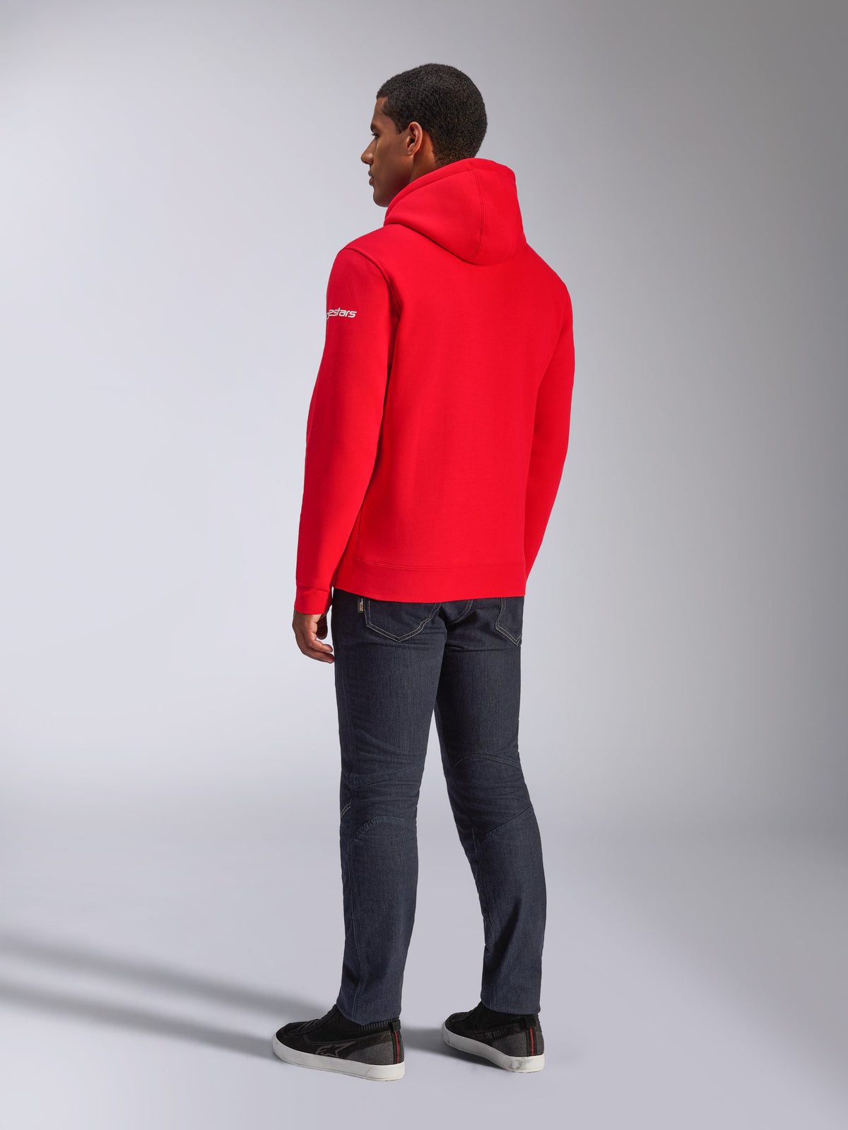 Alpinestars Frontal Hoodie, Red Pullover Fleece Hoodie, back view showing white sleeve wordmark logo, worn with dark denim pants and black shoes, casual motorsport streetwear