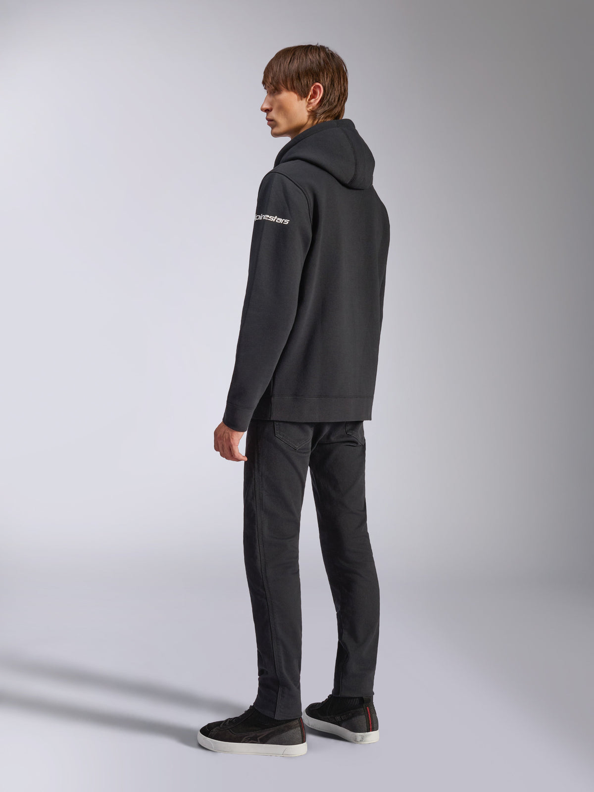 Alpinestars Frontal Hoodie, casual streetwear, black, rear side view showing white wordmark logo on sleeve, worn with black pants and sneakers, modern motorsport-inspired design