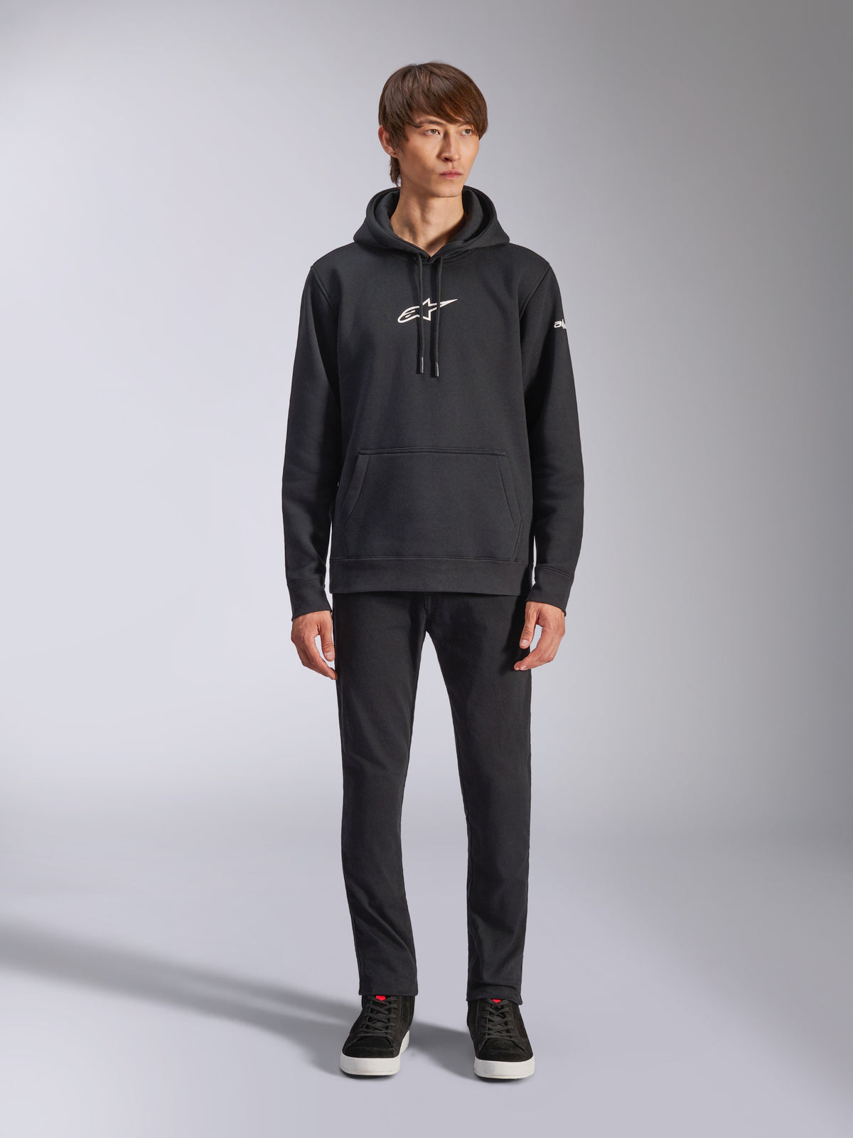 Alpinestars Frontal Hoodie, black pullover fleece hoodie, white Alpinestars logo on chest and sleeve, kangaroo pocket, drawstring hood, worn with black pants, casual motorsport-inspired design