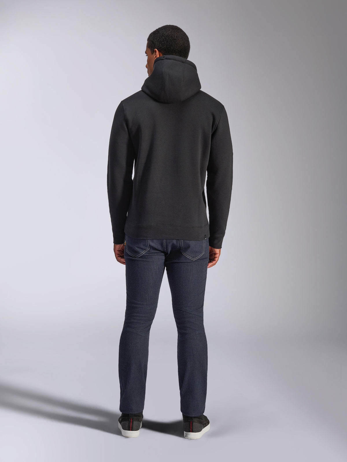 Alpinestars Blaze V3 Hoodie, lässige Streetwear, Schwarz, Rückansicht eines Fleece-Kapuzenpullovers, kombiniert mit dunkelblauer Denim-Jeans und schwarzen Schuhen, cleanes, vom Motorsport inspiriertes Design