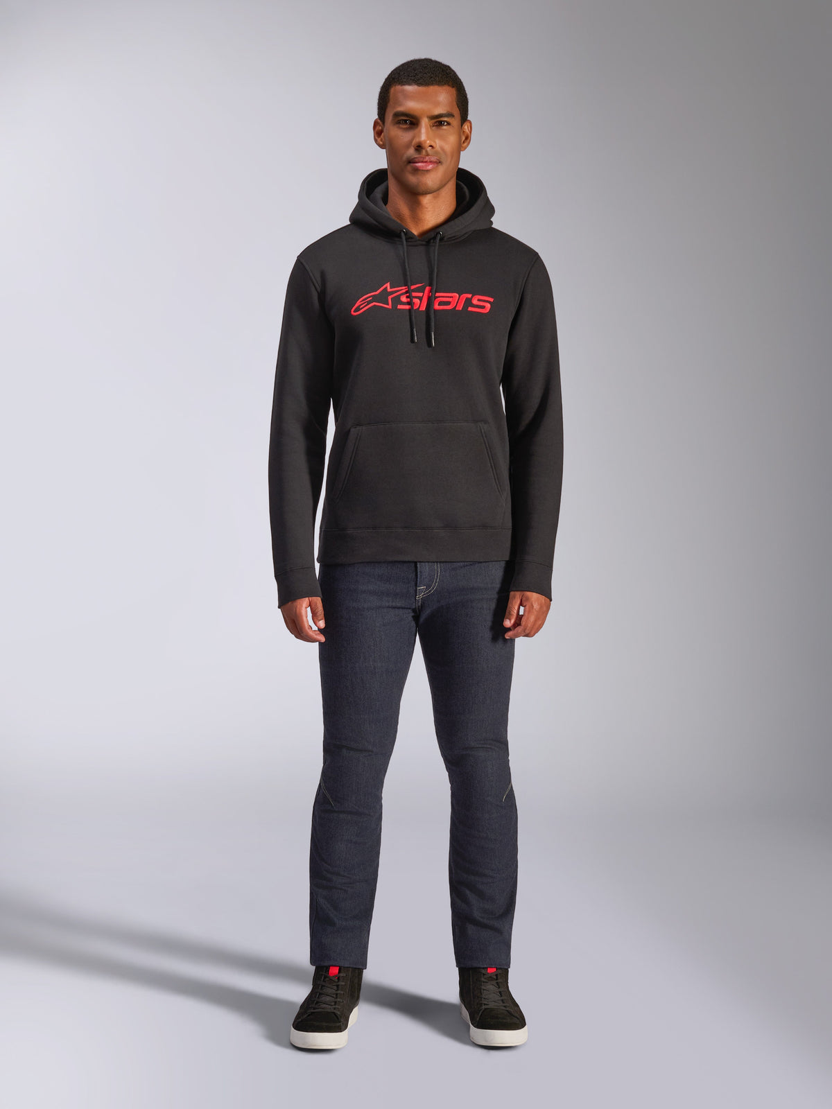 Alpinestars Blaze V3 Hoodie, lässige Streetwear, Schwarz mit rotem Astars-Logo, Vorderansicht, getragen von einem Model mit dunkler Jeanshose und schwarzen Sneakern, modernes, vom Motorsport inspiriertes Fleece-Design